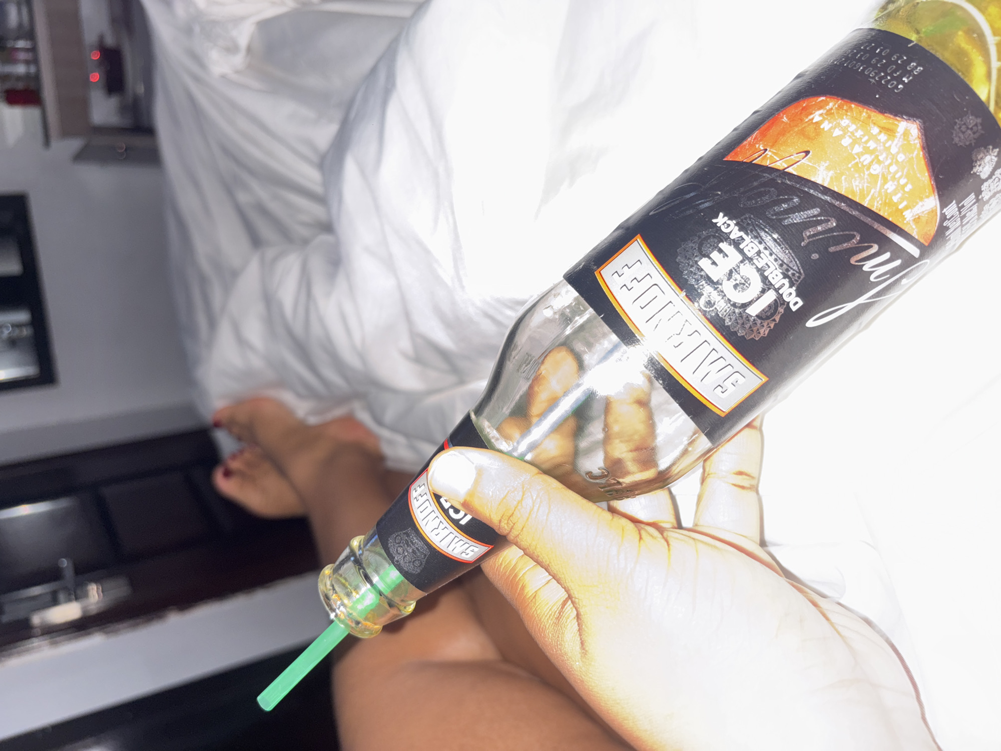 A hand holds a bottle of Smirnoff Ice Double Black, with a green straw inserted in the bottle. The background shows a white bedsheet, a dark-colored cabinet, and a blurred image of bare feet. The location is implied to be in Port Harcourt, Nigeria.