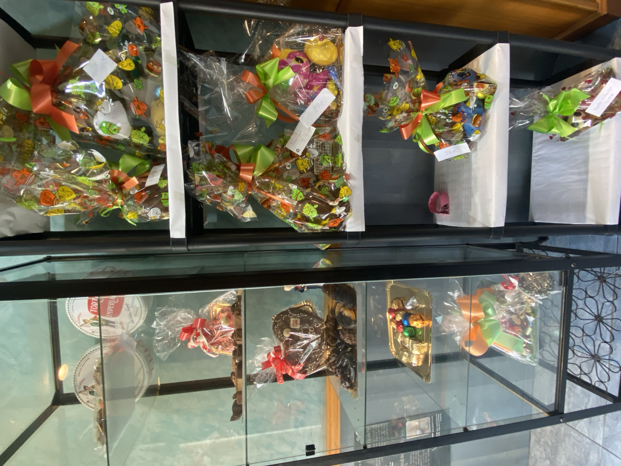 The image depicts a display case in what appears to be a confectionery shop, likely located in Villaviciosa, Spain, given the products and general presentation.
The top shelf contains multiple cellophane-wrapped confectionery items, presumably chocolates or sweets, decorated with colorful bows. The wrappings feature patterns with cartoonish figures and motifs. White dividers separate the arrangements.
The bottom shelf, visible through the glass of the display case, contains additional confectionery items including Torta Imperial boxes, wrapped treats with red ribbons, chocolate heart-shaped sweets, a tray of assorted chocolates, and another cellophane-wrapped treat with orange and green bows.
The overall environment is that of a retail space, designed to showcase and sell sweet treats. The lighting within the display case appears to be moderate, allowing for reasonable visibility of the products.