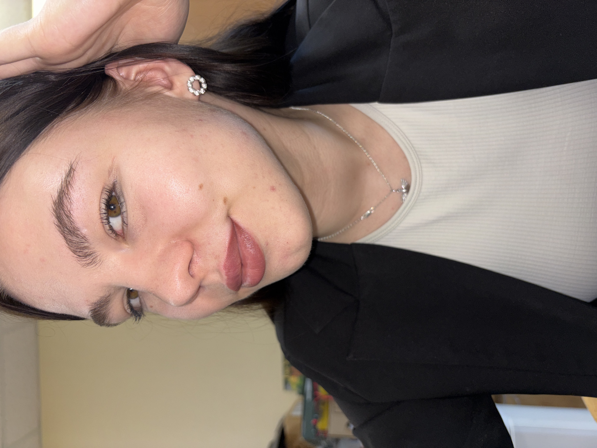 A close-up, rotated portrait of a fair-skinned young woman. She has dark hair, light brown eyes, and is wearing a white ribbed top under a black blazer. She has a silver necklace and floral earrings. The background is a pale yellow wall. The location is not discernible from the image and no evidence is present that would associate the image with Perm, Russia.