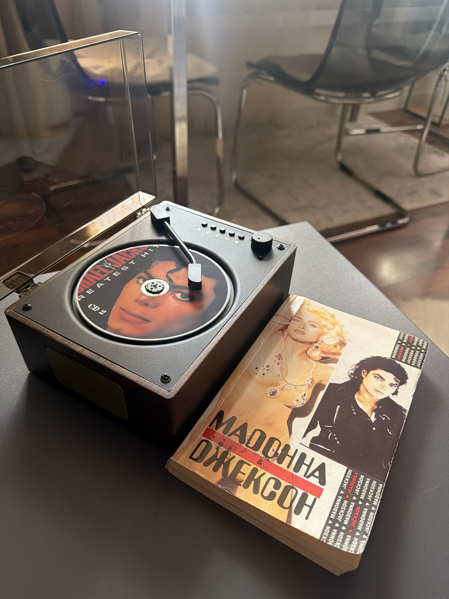 A close-up, overhead shot shows a portable record player with a CD of Michael Jackson's "Greatest Hits" displayed on its turntable. A book with Madonna and Michael Jackson on the cover is placed next to the record player. The record player has a wooden base and a dark gray top. The book is open, showing the spines of the pages. The setting appears to be indoors, with a blurred background of chairs and a wall. No specific location in Otradnaya, Russia, is visible or implied in the image.