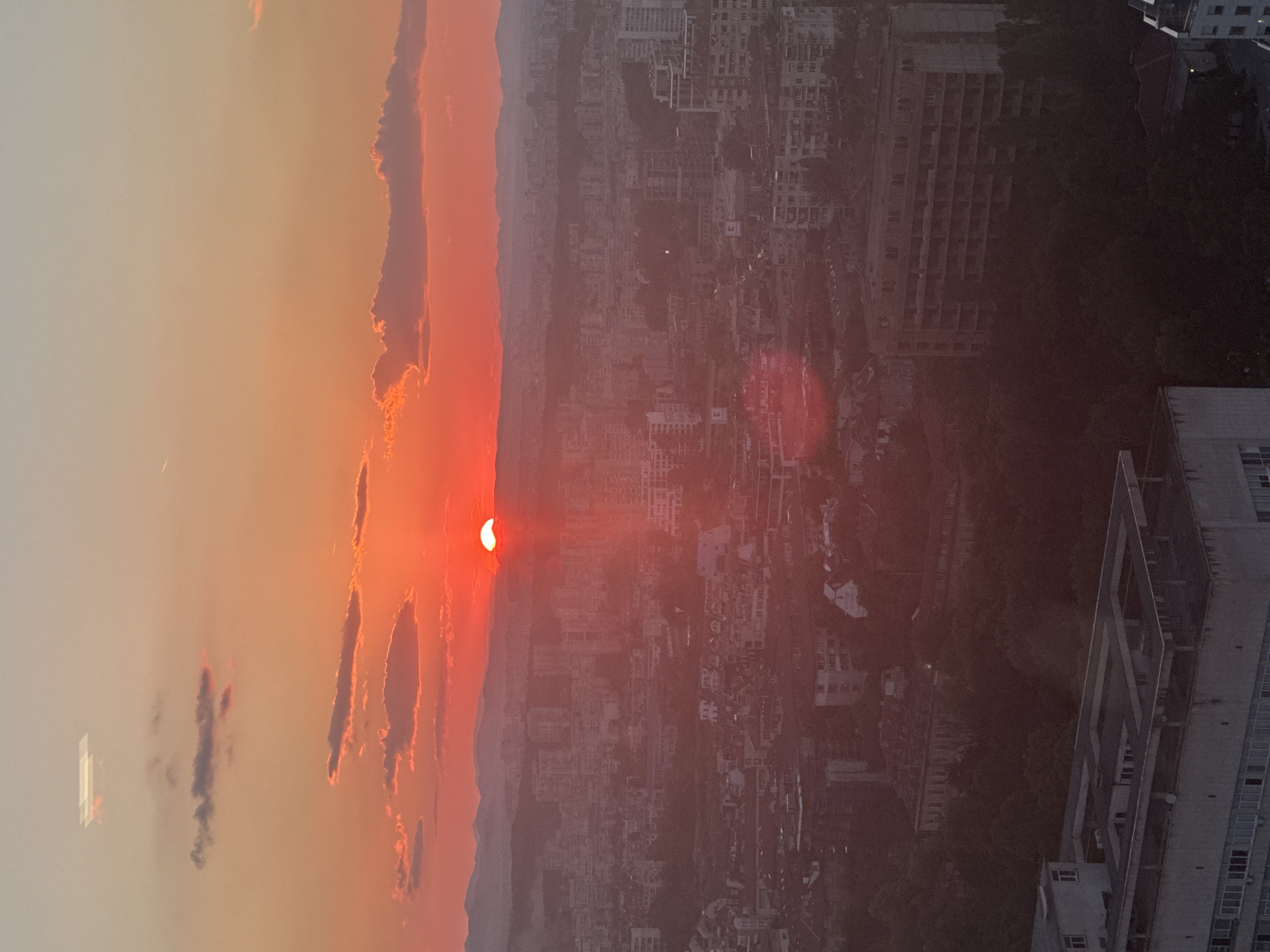 This is an aerial, wide-angle photograph of Sofia, Bulgaria, taken during sunset. The sun, a vibrant red disc, is partially obscured by the horizon and tinged with orange and pink hues, casting a warm glow across the sky. The sky transitions from a bright orange near the horizon to a lighter, pale yellow towards the top. Scattered clouds, also illuminated by the sunset, are visible in the upper portion of the frame.
Below the sky, a sprawling cityscape of Sofia unfolds. Buildings of various sizes and architectural styles, predominantly in muted, earthy tones, are densely packed. Many buildings are multi-story apartment complexes, with some appearing to be under construction or in a state of disrepair, indicated by exposed concrete frames and dark window openings. The city stretches towards a hazy, mountainous horizon in the background, suggesting a vast urban expanse.
In the foreground, a flat-roofed, multi-story building with a visible rooftop structure dominates the lower portion of the image. Other buildings, some with visible windows and balconies, are interspersed with patches of dark green trees, suggesting a mix of urban and natural elements within the city. The overall impression is one of urban density viewed from a high vantage point during a colorful sunset.