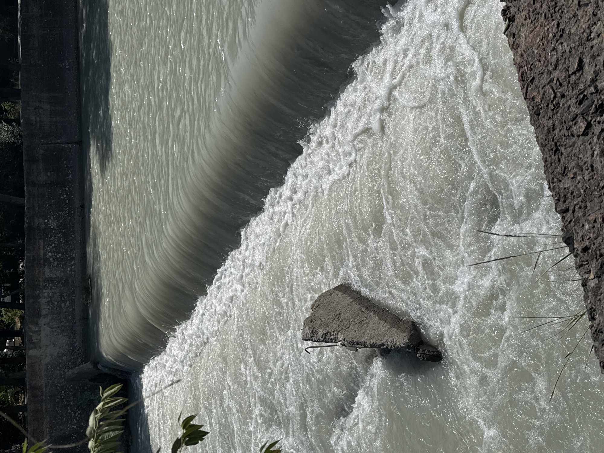 A low-angle, eye-level shot captures a fast-flowing body of water, possibly a canal or river, in Bishkek City, Kyrgyzstan. The water is a murky, light gray color, with white foam and turbulence indicating a rapid current. A concrete barrier, likely a weir or dam, stretches horizontally across the mid-ground, with the water cascading over its edge in a smooth, unbroken sheet. To the left, a large, irregular chunk of concrete with a bent metal rod protruding from it rests partially submerged in the churning water, creating an obstacle that further disrupts the flow.
In the foreground, a rough, dark brown embankment is visible, composed of jagged rocks and soil. Some dry, thin blades of grass sprout from this embankment. The background features a dark, textured concrete wall, likely part of the canal or embankment construction. Above the wall, a dense line of dark green foliage from trees forms a natural canopy, suggesting a park or wooded area adjacent to the water. Sunlight glints off the water's surface in various spots, creating a dappled effect. The overall impression is one of a managed urban waterway with signs of wear and tear.
