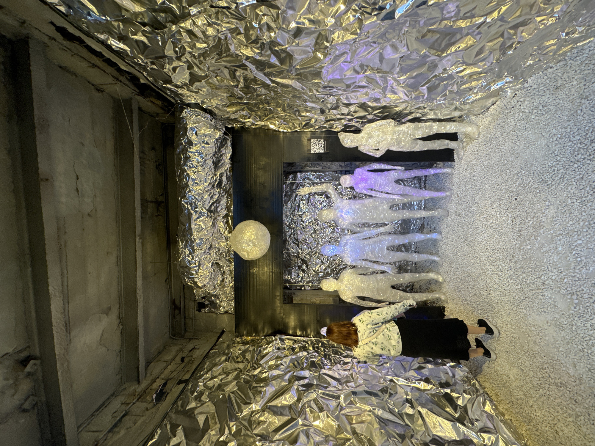In Saint Petersburg, Russia, within what appears to be an art installation or exhibition space, a person stands facing away from the camera. The individual is wearing a light-colored patterned jacket over a dark skirt and black shoes. They are positioned in a hallway-like area, with the walls and ceiling covered in crinkled, reflective metallic material, resembling aluminum foil. The floor is covered in small, white pebbles.
In front of the person, arranged in a row, are six translucent, humanoid figures. These figures are illuminated from within, with some emitting a white light and others a soft purple glow. One figure on the far right is more opaque and appears to be made of a similar translucent material.
Above the figures, suspended from the ceiling, is a spherical, textured light fixture. The overall ambiance suggests a contemporary art display with a focus on light and reflective surfaces. The exposed concrete elements on the ceiling and walls, along with the metallic covering, contribute to an industrial or raw aesthetic.
