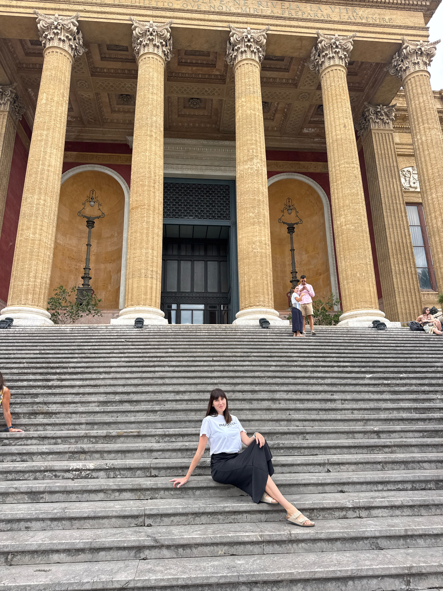 The image shows the grand staircase leading to the Teatro Massimo in Palermo, Italy. A woman in a white t-shirt and black skirt is sitting on the stairs, posing for the camera. In the background, a couple is standing on the stairs, seemingly taking a photo. The architecture of the Teatro Massimo, with its imposing columns and ornate details, is visible behind them. Other people are scattered on the stairs, some sitting and others walking. The overall scene depicts a vibrant and lively atmosphere at one of Palermo's most iconic landmarks.