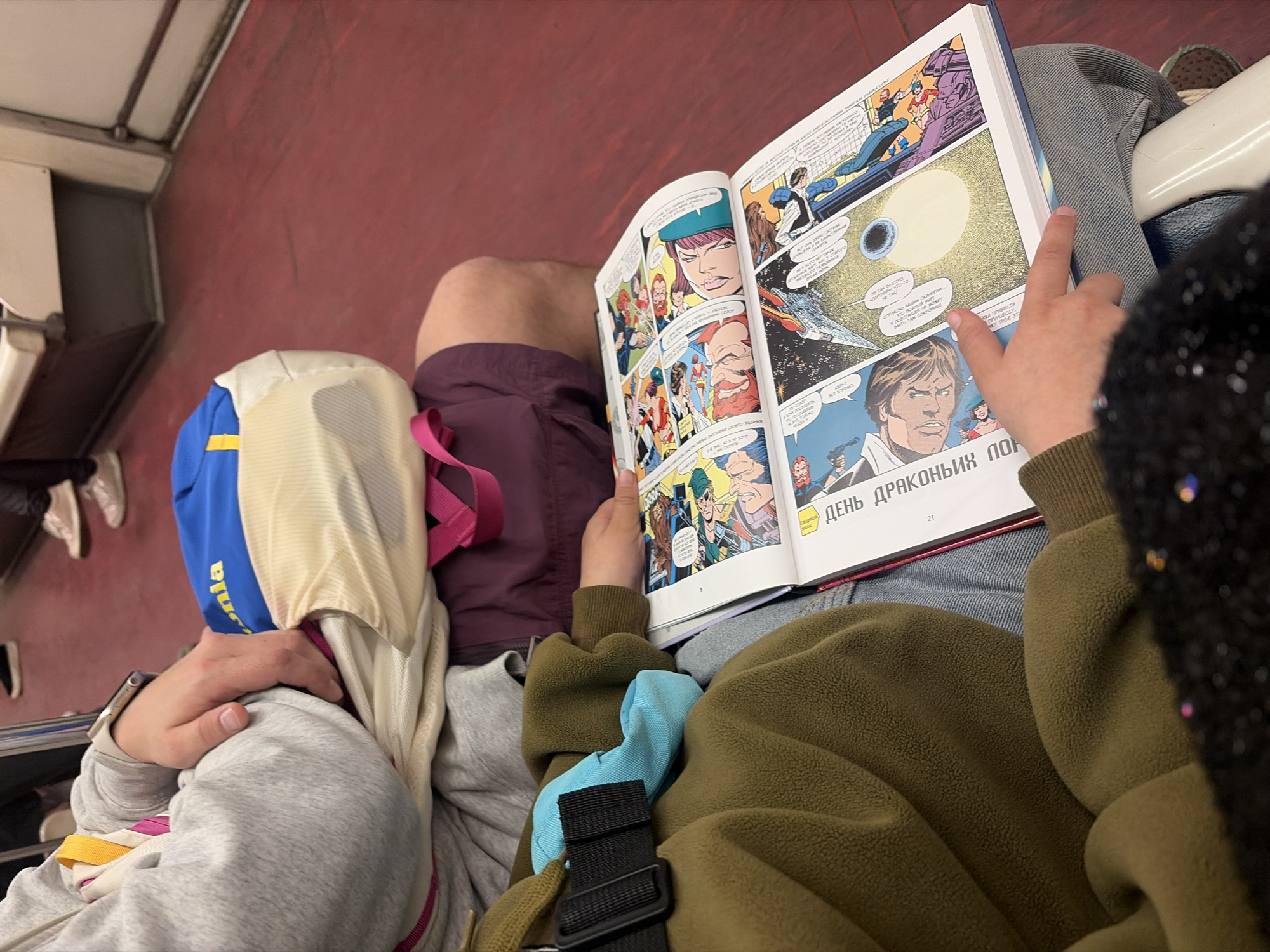 Two individuals are seated next to each other, presumably on a train. The background appears to be the interior of a train carriage with a red-brown wall and luggage rack. One person is engrossed in reading a large, open comic book, while the other person is leaning back with their head covered, possibly asleep. The comic book appears to be written in Russian, and the page shown features an illustration of a solar object. One person is wearing an olive-green fleece jacket and denim jeans. The other person is dressed in a grey hoodie. The scene takes place in Saint Petersburg, Russia.