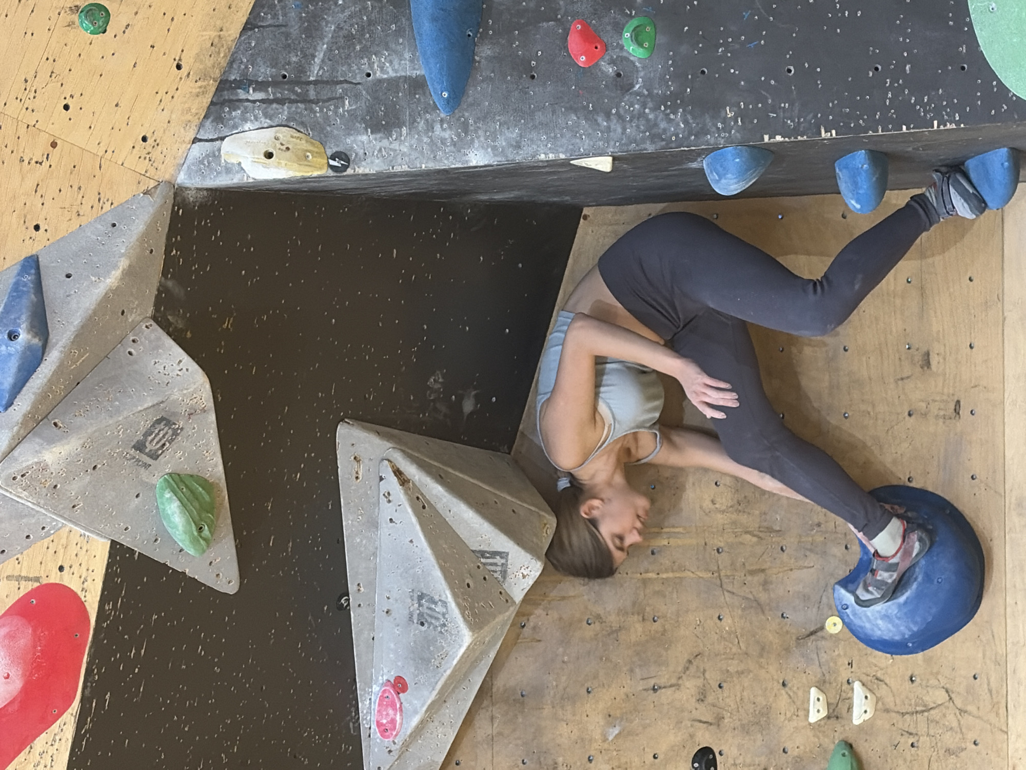 The image shows a young woman indoor rock climbing. She is wearing a light blue sports bra and dark leggings. Her right foot is placed on a large blue climbing hold, and her left foot is on a smaller blue hold. She is looking down, her head tilted as she positions herself on the wall. The climbing wall is made of wood panels and features various colored climbing holds, including red, green, yellow, and blue. There are also large, angular grey structures attached to the wall, providing different angles and challenges for climbers. The lighting is artificial and appears to be from overhead. The background is a dark, textured surface. The scene appears to be in an indoor climbing gym. There is no indication of the location being Moscow, Russia, in the image itself.