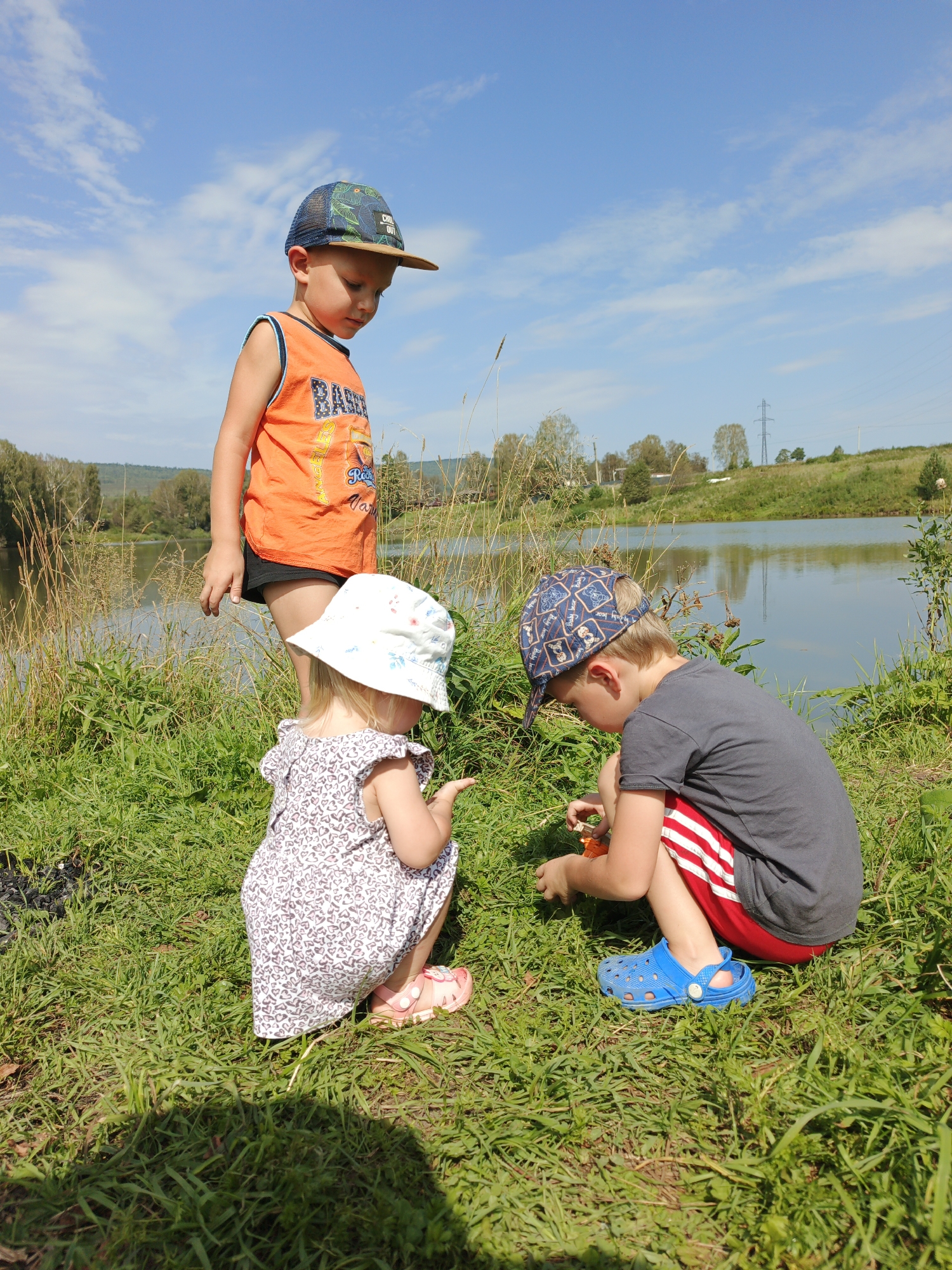 Three young children are on the grassy bank of a lake on a sunny day. The child on the left, a boy wearing an orange sleeveless shirt and a patterned baseball cap, stands looking down. In the center, a toddler girl wearing a white sun hat and a floral dress kneels, looking at something on the ground. To the right, a boy in a grey t-shirt, red shorts with white stripes, and a blue baseball cap kneels and appears to be playing with something orange in his hands. The lake is calm and reflects the blue sky with scattered white clouds. In the background, there are trees and rolling hills. The location is identified as Urtiushka, Russia.