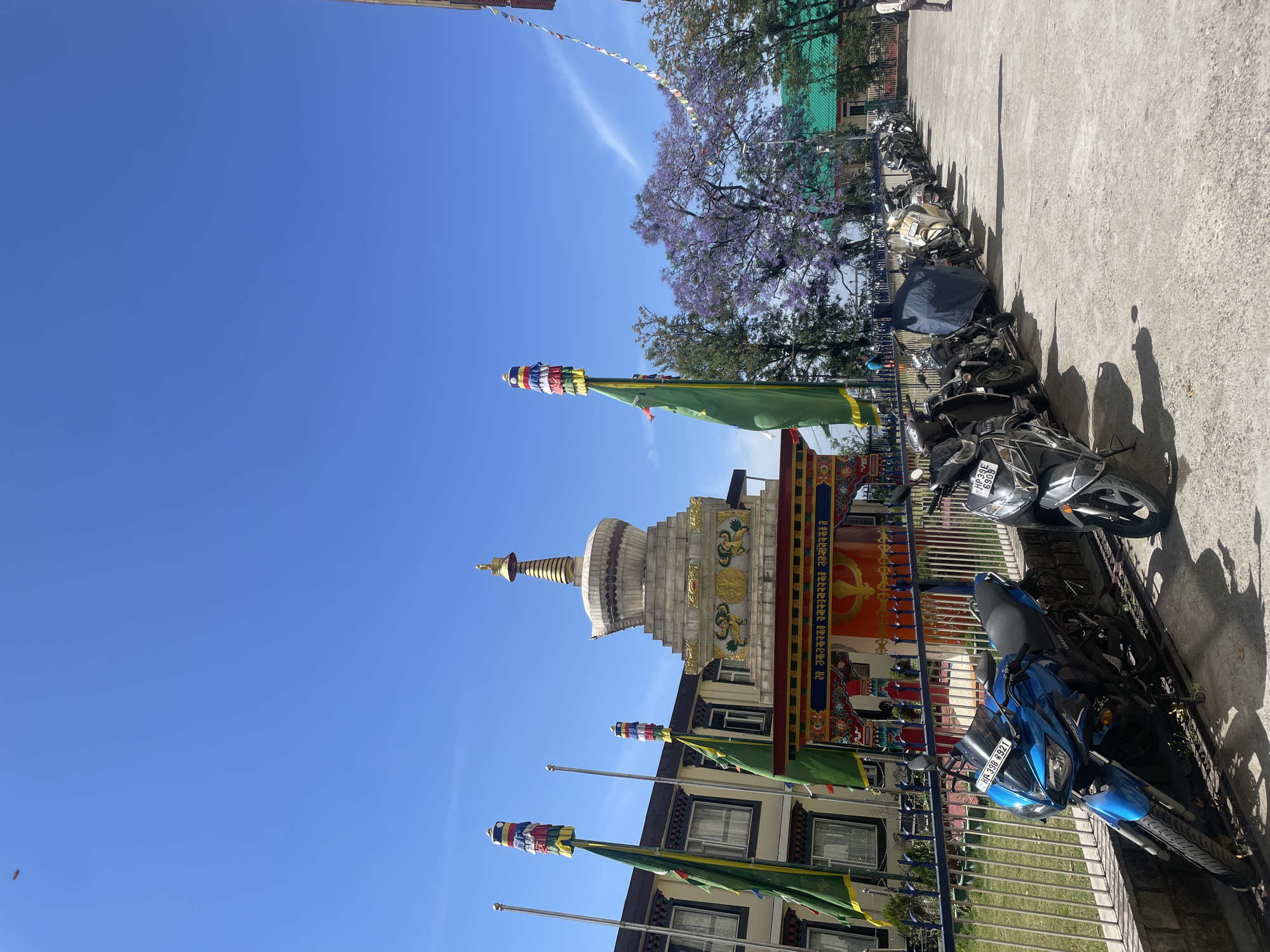 The image depicts a Buddhist stupa in Dharamshala, India, under a clear blue sky. Several motorcycles are parked on the right. A flowering tree with purple blossoms is visible in the background. Prayer flags are attached to poles in front of the building. A metal fence separates the building from the parking area.