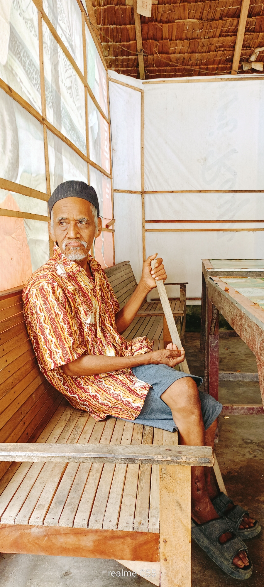 An elderly man with a gray beard and a black cap is sitting on a wooden bench. He is wearing a patterned shirt and blue shorts, and holding a wooden stick. The man is sitting under a thatched roof structure with white tarpaulin walls. To the right, there is a wooden table with what appears to be a patterned surface. The overall environment suggests an outdoor or semi-outdoor setting, possibly in a rural area. The image was taken in Lhokseumawe, Indonesia, judging by the "realme C55" watermark, which is a model of smartphone produced by Oppo, a company that has a significant presence in Southeast Asia.