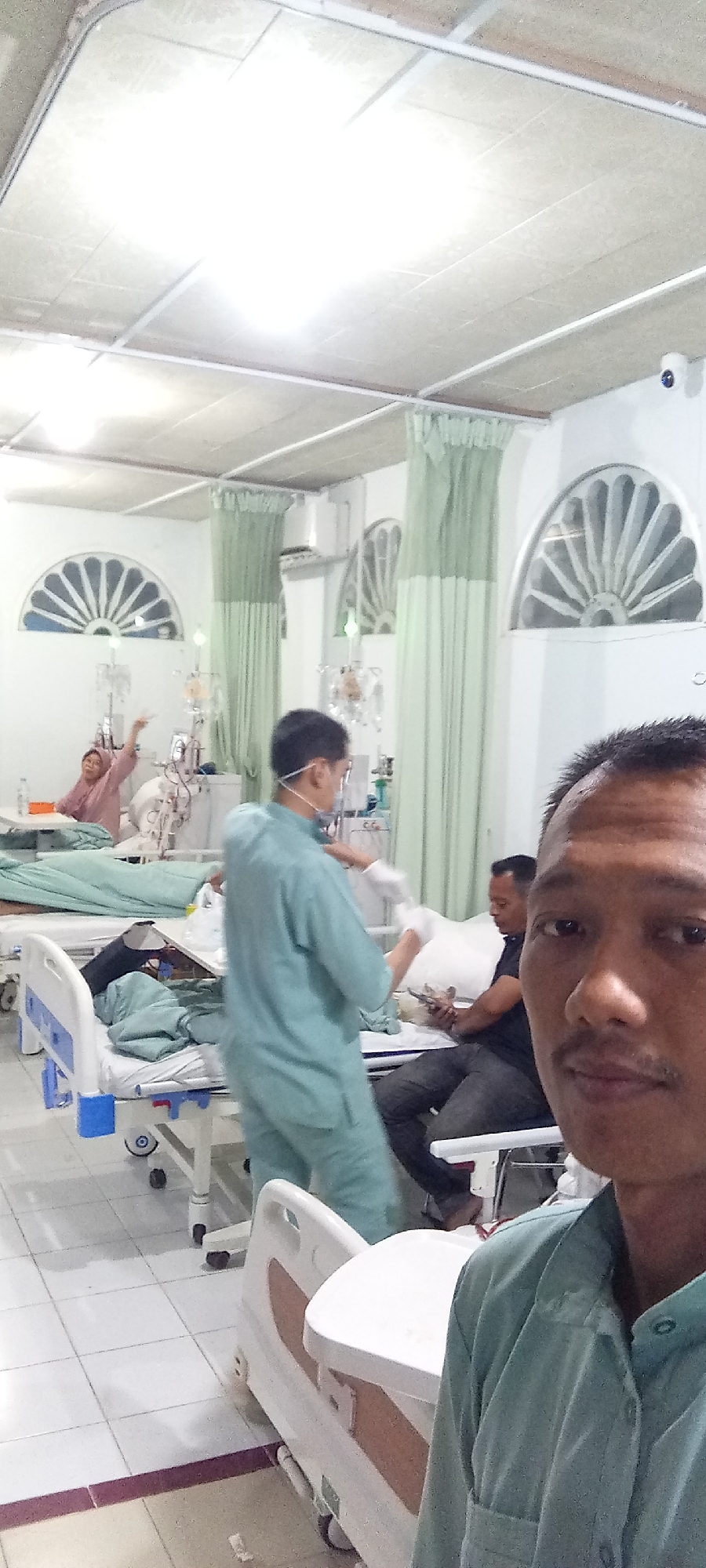 The image shows an indoor scene in Lhokseumawe, Indonesia, appearing to be a hospital room. The room has multiple hospital beds, and several people are visible. One person, wearing a light green scrub suit, is attending to a patient lying in a bed. Another patient in a headscarf is in bed on the far left. A man in dark clothing is seated by a bed. The person taking the photo is in the foreground wearing a similar light green scrub suit. The room is well-lit with a tiled floor and curtains beside arched windows. Medical equipment is visible, suggesting a healthcare setting.