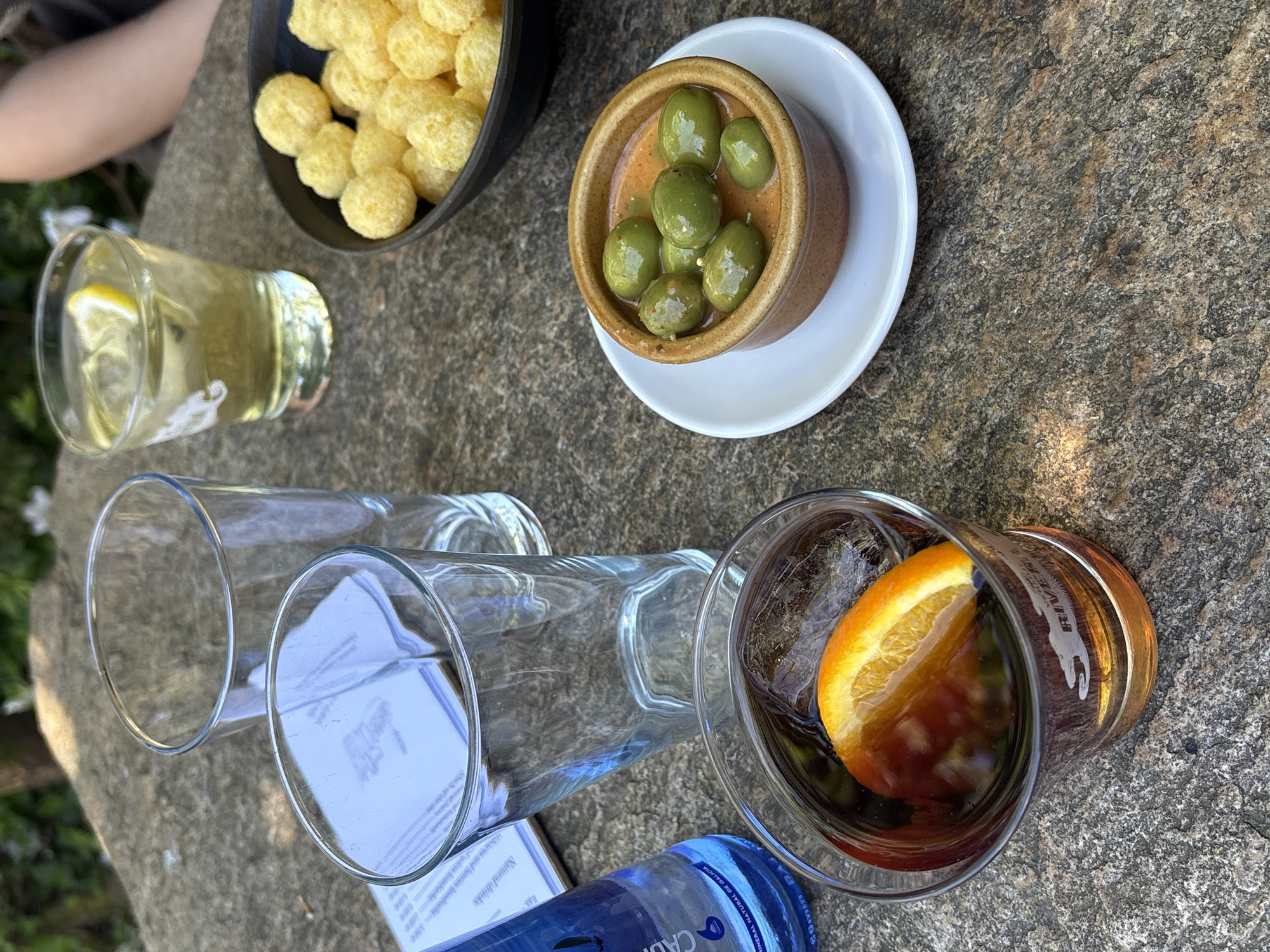 The scene shows a collection of refreshments and snacks arranged on a weathered stone surface, likely an outdoor table. Clockwise from top left: A bowl filled with cheesy puff snacks, a small dish of green olives in a light-colored sauce on a white saucer, a glass containing a dark liquid with an orange slice, two empty glasses lying on their sides, a bottle of water, and a glass with a light-colored liquid and a lemon slice. A menu card is partially visible under one of the empty glasses. Part of a person's arm is visible in the upper left. The presence of tapas and drinks indicates a casual gathering, potentially a social occasion. The location is Santiago De Compostela, Spain, evident from the bottle of "Cabreiroa Natural Mineral Water" which has "Natural De Galicia" listed on the bottle.