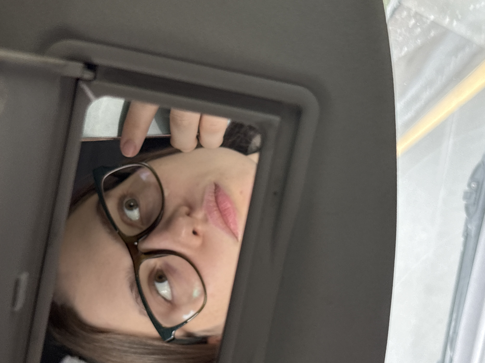 The image depicts a person with brown hair and glasses reflected in a car's sun visor mirror. The person's face is partially obscured by the mirror frame and their own finger. Outside the car window, the background is blurred, obscuring detail. The image does not provide any information regarding the location in Myaglovo, Russia.