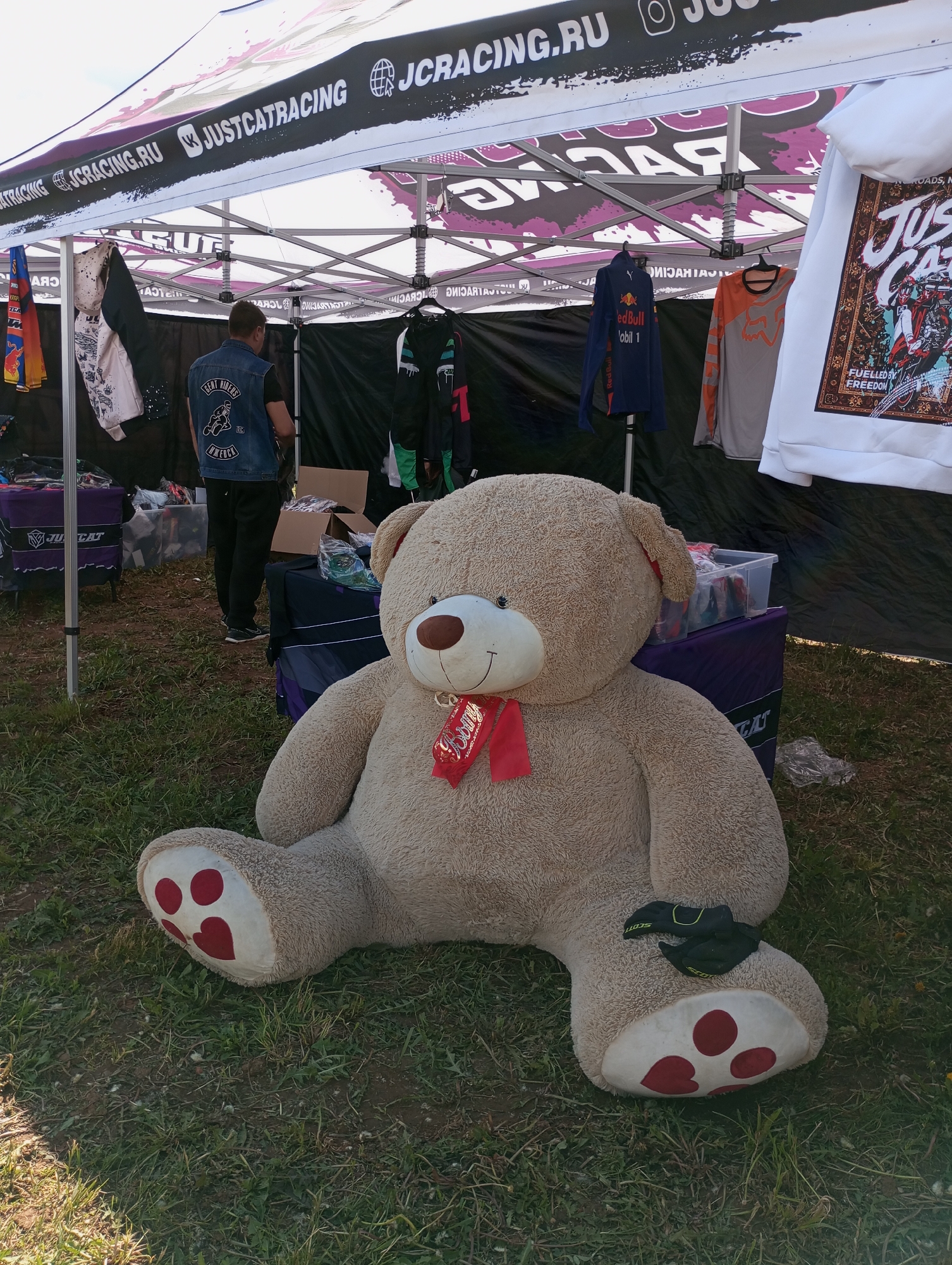 The image shows a large, tan teddy bear with a red ribbon around its neck, sitting on grass. It has a white paw with three red heart-shaped markings. On the paw, there's a pair of black gloves. Behind the teddy bear, there's a tent set up with the words "JUSTCATRACING JCRACING.RU" printed on it. There are clothes hanging inside the tent, including a Red Bull-branded shirt and a white hoodie. A man wearing a denim vest is standing inside the tent. Two tables filled with clothes are visible inside the tent. The location could be Izhevsk, Russia, based on the "JCRACING.RU" domain, but there's no definitive evidence to confirm it.