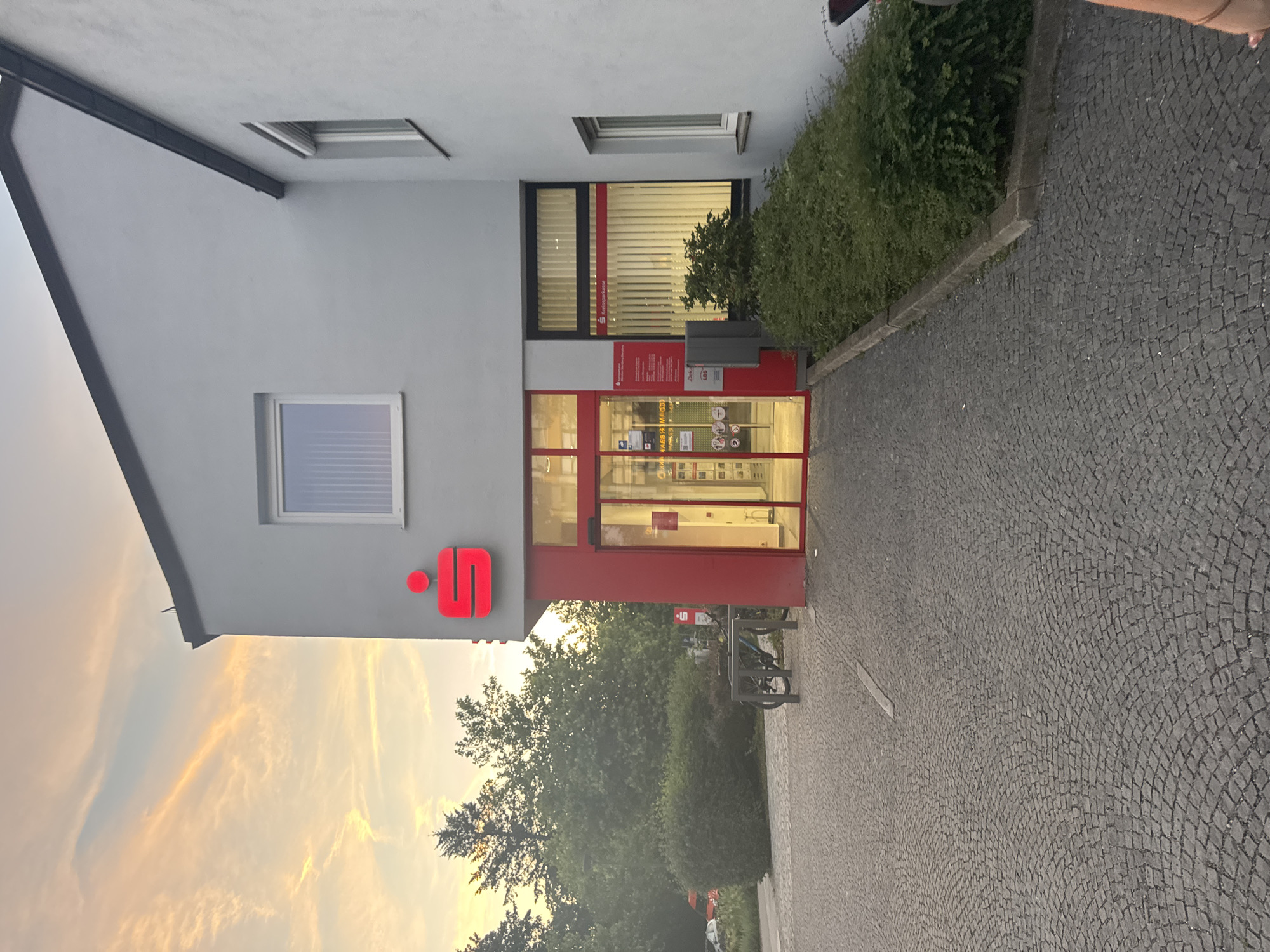 The image shows the exterior of a Sparkasse bank branch in Ottobrunn, Germany. The building is light gray with a red entrance area and a prominent red Sparkasse logo. The sky above displays a sunset with colorful clouds. In front of the bank, there is a cobblestone pavement and a bicycle rack with two bicycles. Green bushes and trees are visible in the background. The scene appears to be calm and quiet, with no people actively engaged in any transactions. The lighting suggests it is late afternoon or early evening during sunset.