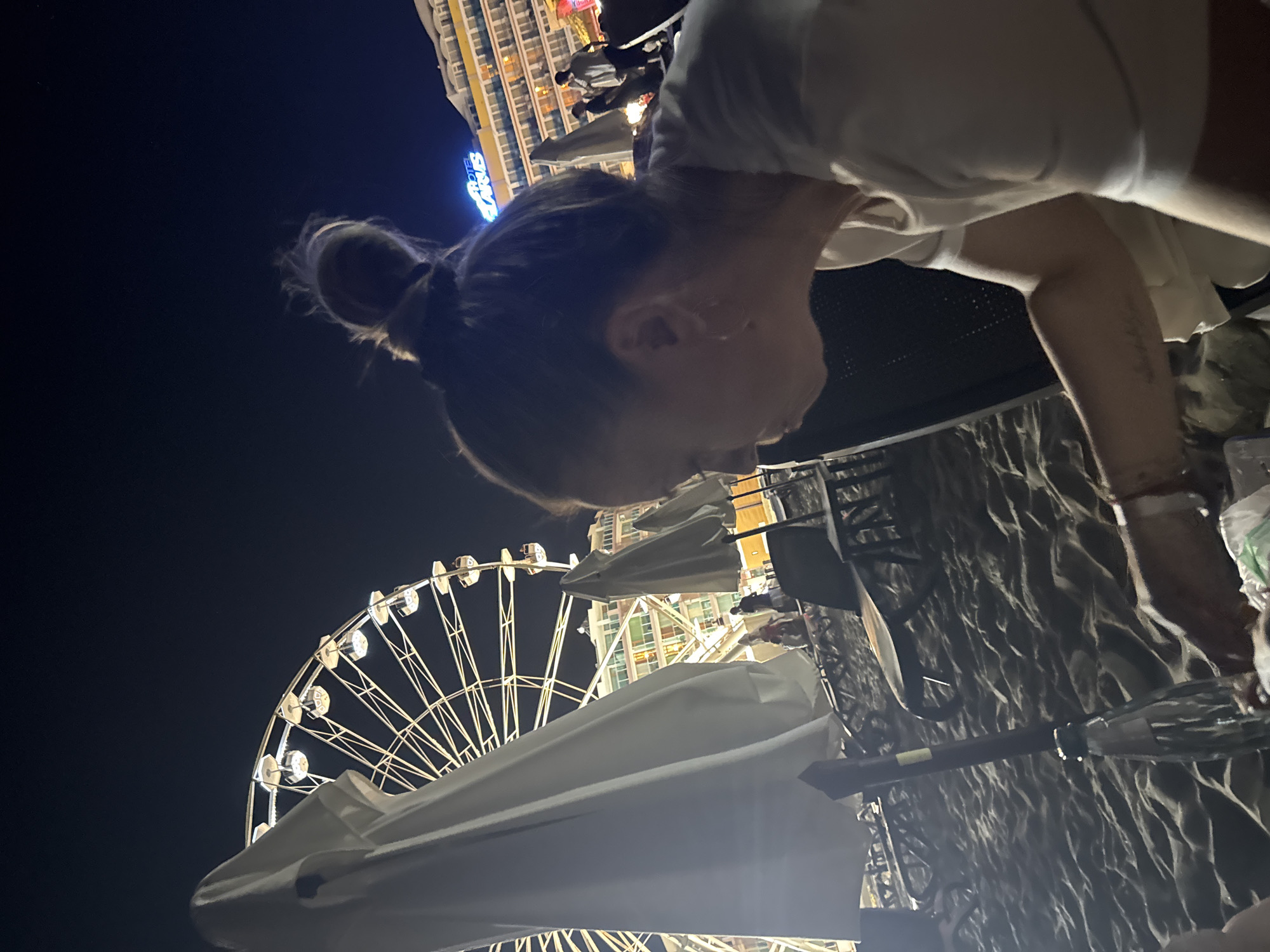 In Nesebar, Bulgaria, at night, a woman with her hair in a bun is positioned in the foreground, her right side facing the camera. She is wearing a white t-shirt, and a tattoo is visible on her left forearm. To her left, a large white beach umbrella dominates the frame. In the background, a brightly lit Ferris wheel is prominent against the dark sky, and a hotel building with illuminated windows is visible. The scene is set on a sandy beach, with beach chairs and other umbrellas scattered around. The overall impression is a nighttime scene at a seaside resort.
