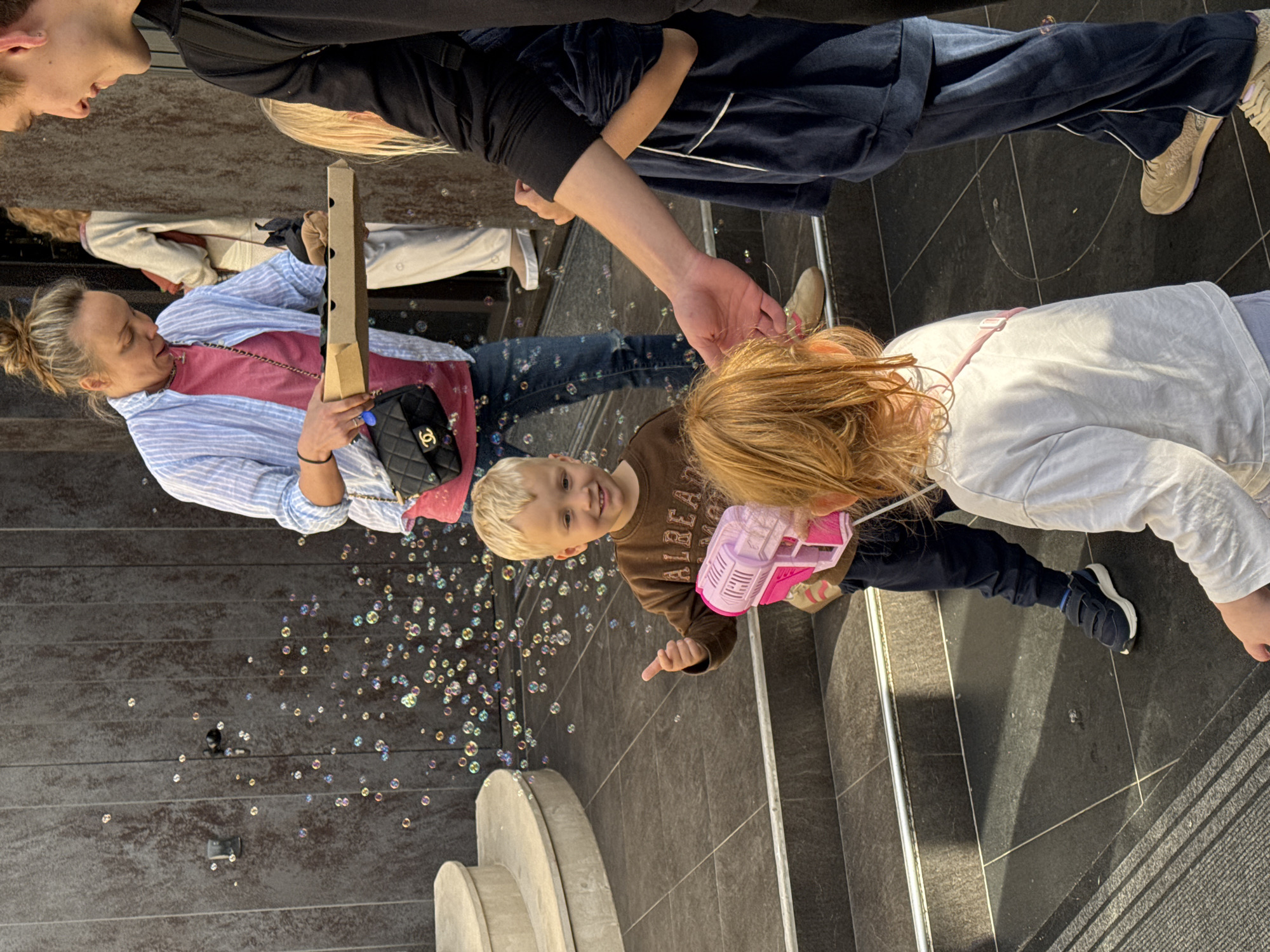 The scene captures a group of people standing on a stone tile surface, likely a sidewalk or plaza. A young blonde boy in a brown shirt points at the camera while holding a pink bubble gun which has sprayed a multitude of bubbles into the air. To his left, a woman with her hair in a bun is holding a triangular piece of cardboard and a small black purse, a blue and white striped shirt over her pink top. An adult’s arm is partially visible. Further back, another person wearing light-colored trousers and a white shirt is partially seen. In the top of the image, another adult in dark clothing looks down at the group, with another person, also in dark clothing, visible next to them. The backdrop is a dark, textured wall. The location is Budva, Montenegro.
