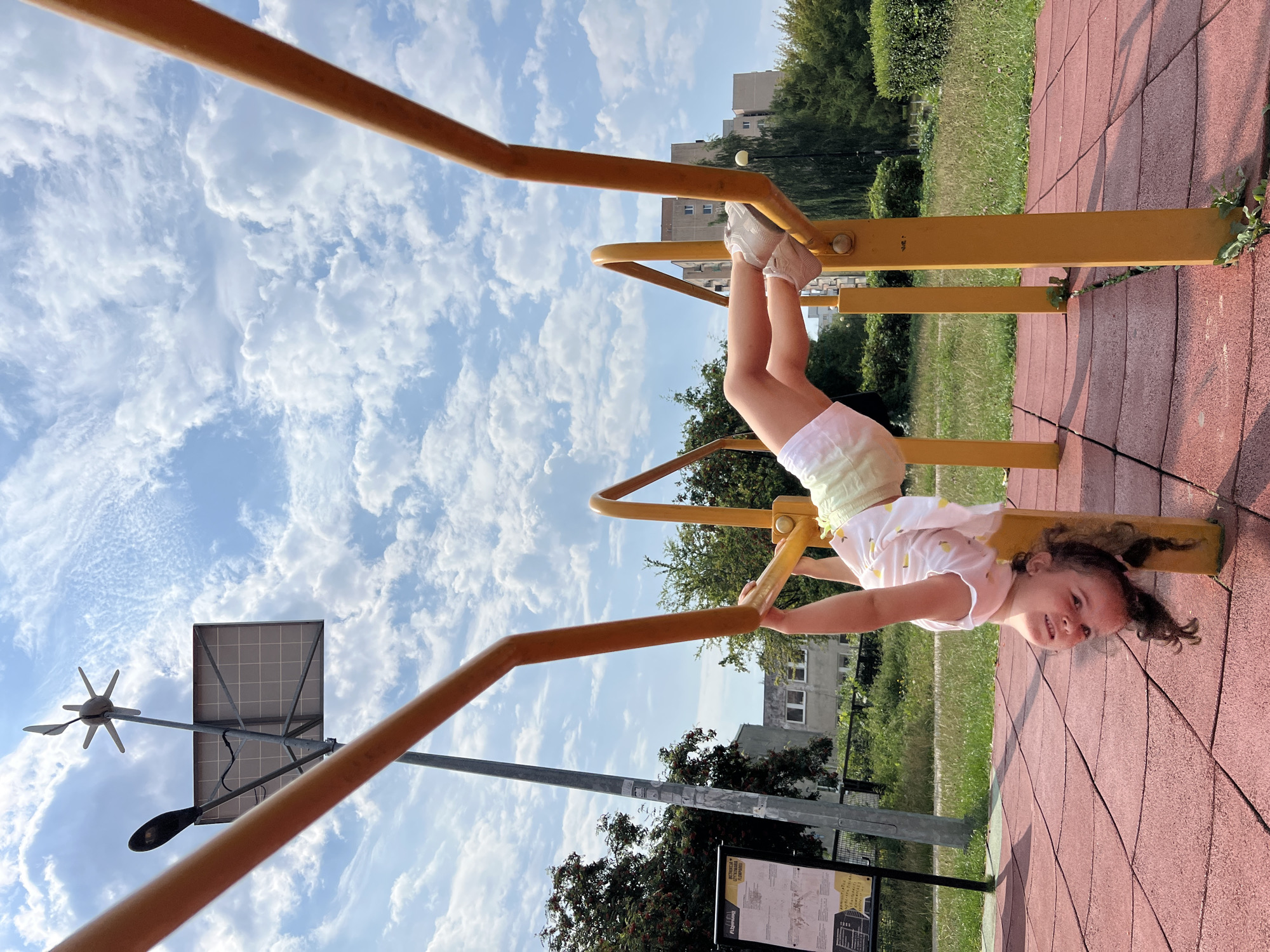 A young girl is hanging upside down from yellow monkey bars in an outdoor park in Lublin, Poland. She is wearing a white t-shirt with a lemon print and light-colored shorts. Her sneakers are visible on the bars. The ground is covered with red rubber safety tiles. In the background, there is green grass, trees, and apartment buildings. Above, the sky is blue with white, fluffy clouds. A solar panel and a small wind turbine are visible on a pole to the left of the monkey bars. A sign with instructions is also mounted on a pole in the left background.