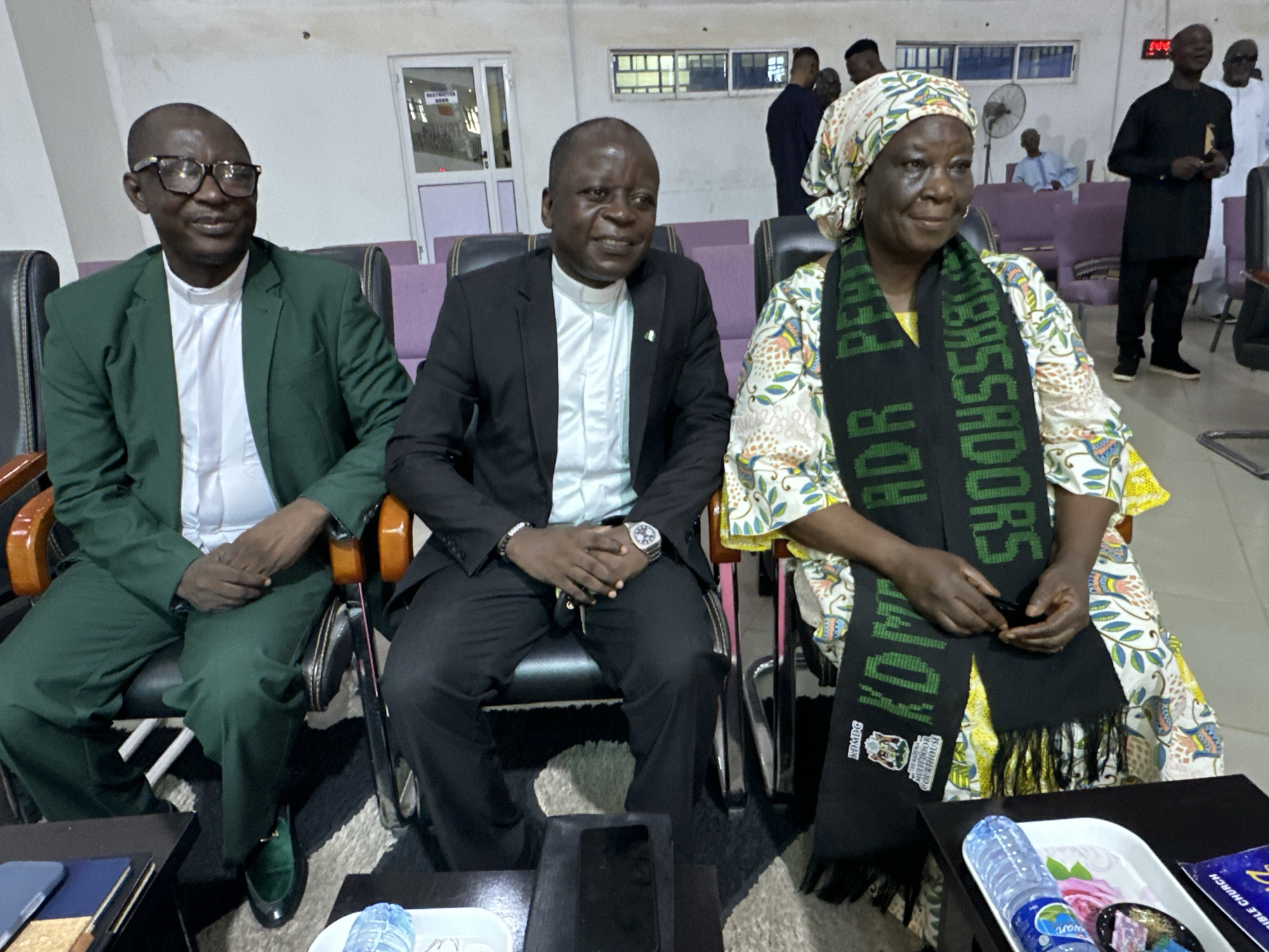 Three individuals are seated in chairs in what appears to be an indoor event space. On the left, a man wearing glasses and a green suit with a white collar shirt is seated. In the center, a man in a black suit and white collar shirt is seated. To the right, a woman wearing a headscarf and a colorful patterned dress with a black scarf around her neck is seated. The woman's scarf has green lettering and a crest. In the background, other individuals are visible, some standing and some seated. The environment features chairs, tables, a fan, and doors. This scene is located in Goni Gara, Nigeria.
