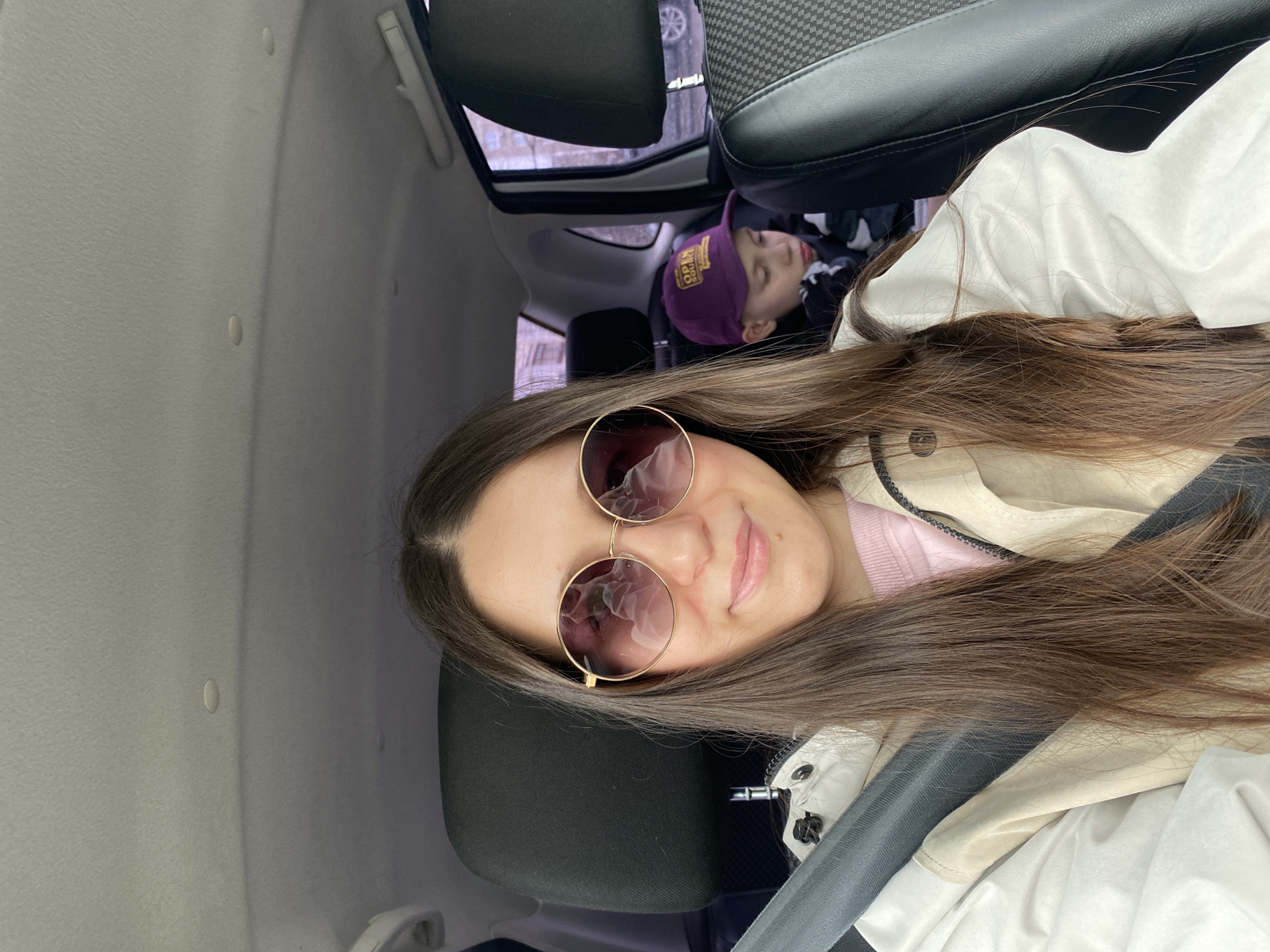 The image appears to be taken inside a car, possibly a selfie. A woman with long brown hair and sunglasses is in the foreground, wearing a light-colored jacket over a pink sweater. In the background, behind the woman's seat, a child wearing a purple hat is asleep. The car interior is visible, including the seats and headliner. Based on the context provided, the location might be Chaikovsky, Russia, but this is only a speculative element of the scene, as no specific evidence about the location is provided.