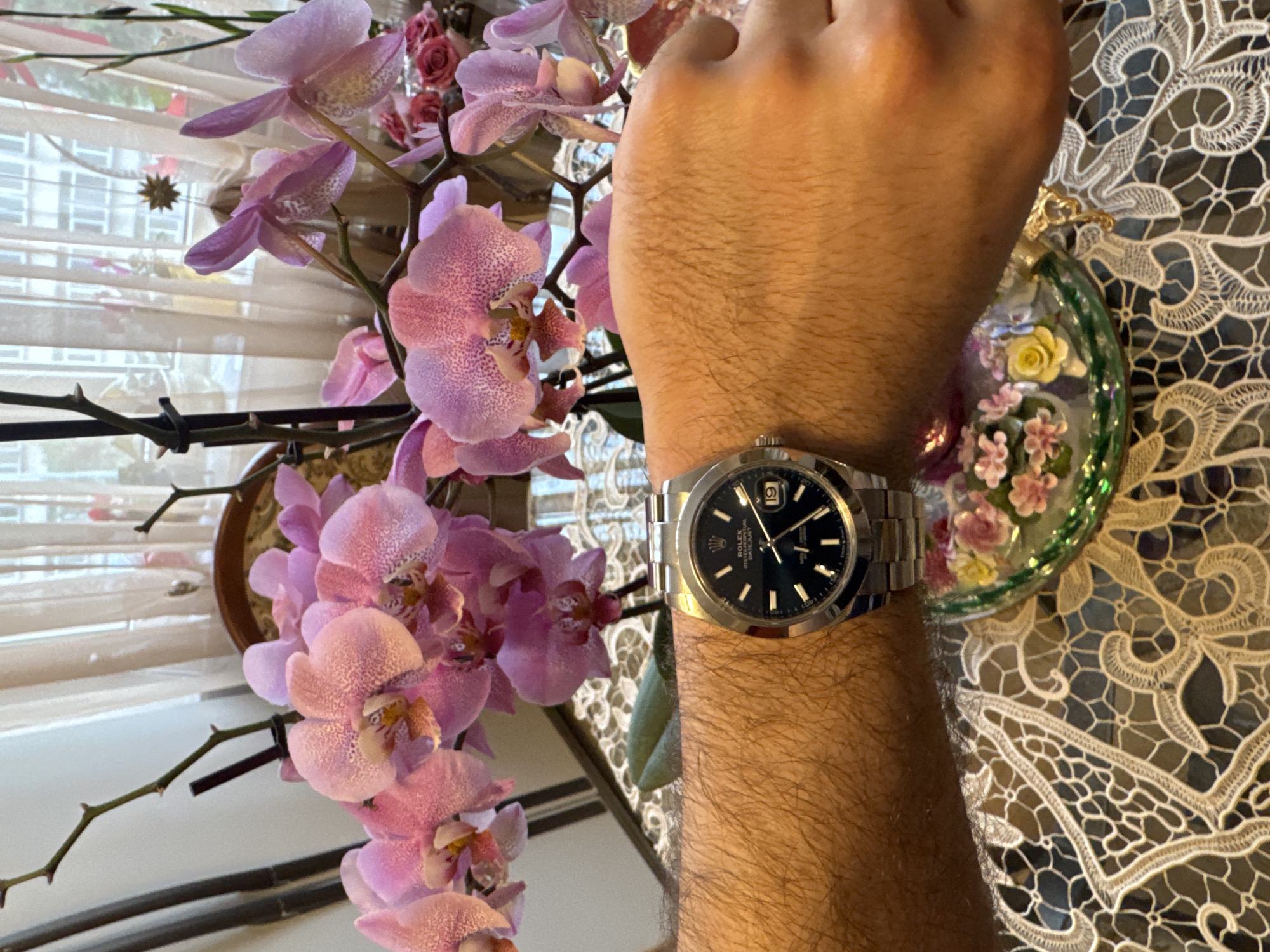A photograph shows a person's arm with a Rolex Datejust wristwatch. The watch has a dark blue dial, stainless steel case and bracelet, and a date window at the 3 o'clock position. The arm is hairy. The background contains a plant with pink orchids, a window with blinds, and a decorative lace tablecloth. A floral ornament hangs from the tablecloth. The location is not evident from the image. It is not possible to tell if this image was taken in Plovdiv, Bulgaria.