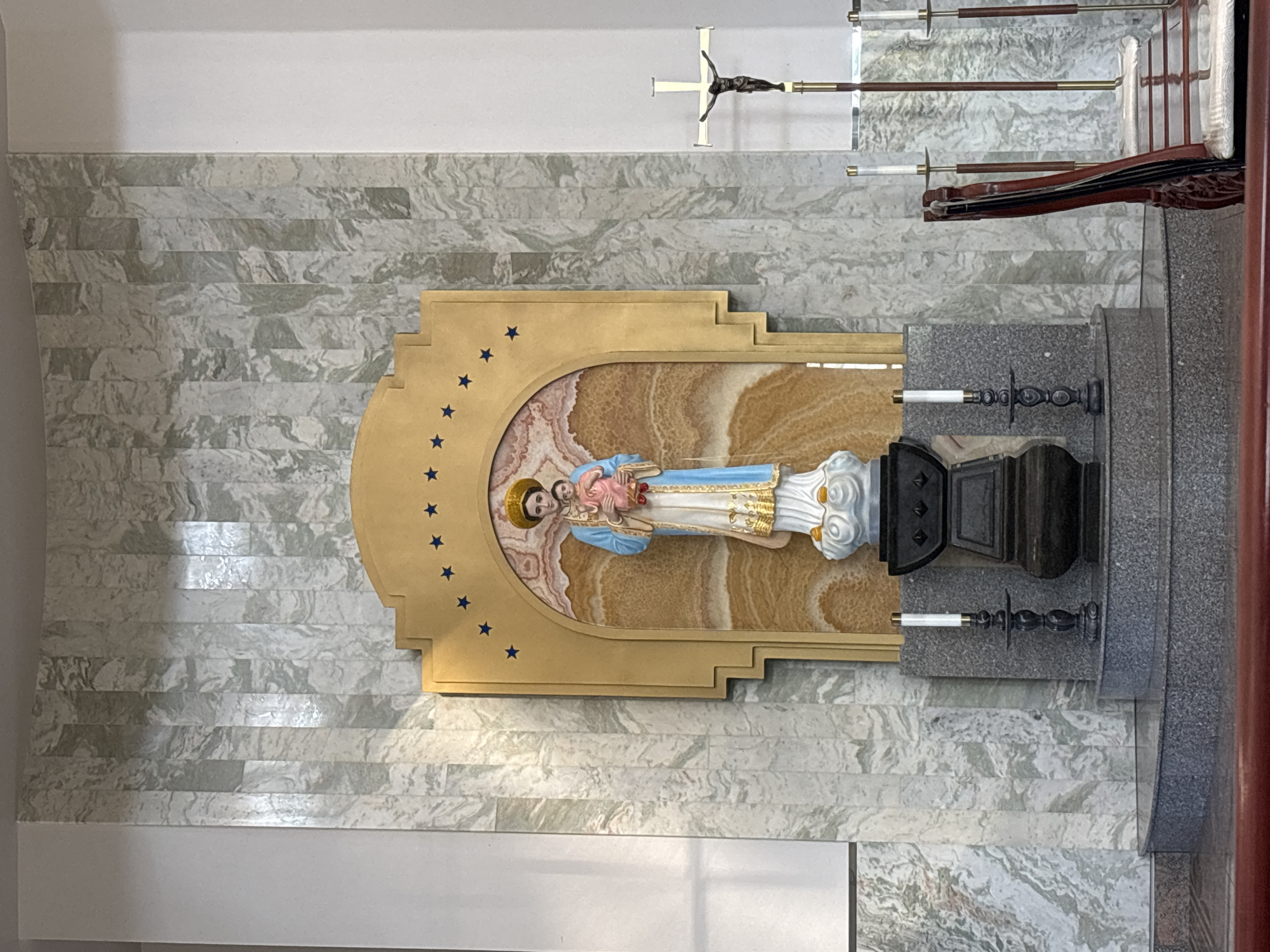 The image depicts a religious altar within a building, likely a church, in Phoenix, United States of America. A statue of a woman holding a baby is centrally positioned on a multi-tiered, grey, speckled platform. The statue is placed within an arched alcove adorned with a golden frame and blue stars. To the right of the statue, a crucifix stands tall. Two candlesticks are visible on the platform, one to the left and another slightly behind and to the right of the central statue. The wall behind the altar features a decorative marble pattern. In the foreground, a dark red wooden railing and seating area are partially visible.