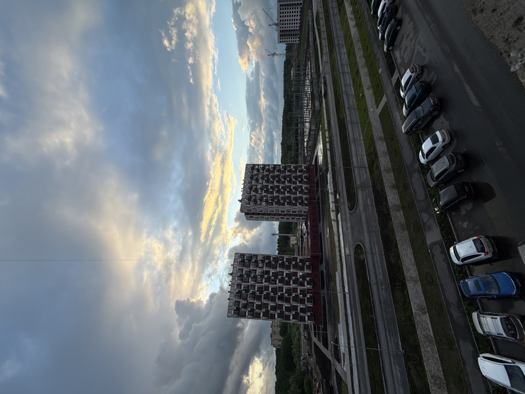 The image shows a view from a high vantage point, possibly a tall building, in a city assumed to be Veliky Novgorod, Russia. The sky is prominent, filled with a mix of dark, heavy clouds on the left and lighter, sunlit clouds towards the right, indicating varying weather conditions. Below the sky, two tall, modern apartment buildings are visible, characterized by a grid-like pattern of windows and balconies. These buildings are positioned close to each other, suggesting a dense urban development. Adjacent to the buildings is a multi-lane road with parked cars along the side. The road is wet, indicating recent rain. Beyond the road, there is a green area with trees. In the distance, construction cranes and the skeleton of another building suggest ongoing urban development.