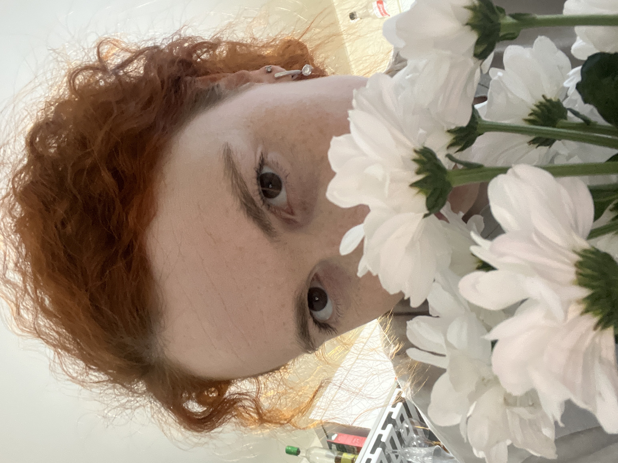 A close-up, tilted, eye-level shot depicts a fair-skinned person with freckles and reddish-brown, curly hair. The person's face occupies the left side of the image, with their brown eyes looking upwards. A small, metal piercing is visible on the upper part of their ear. To the right of the person's face is a bouquet of white flowers with visible green stems and dark green centers. Below the flowers and the person's face, a white surface is visible with various objects including a white plastic basket, bottles of liquid with green caps, and other blurred items. No specific landmarks or features are visible to confirm the location as Chaikovsky, Russia.