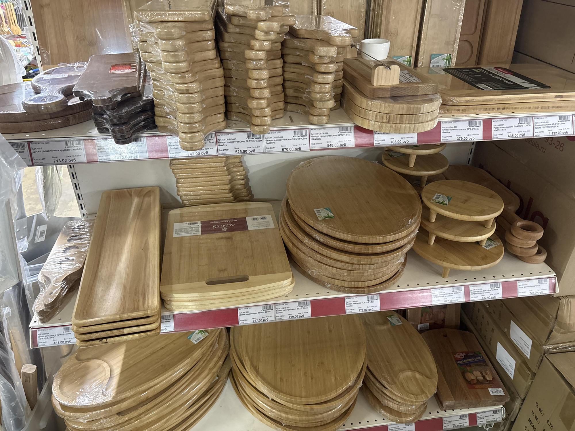 This is an indoor shot of a retail display showcasing a variety of bamboo cutting boards and serving platters. The scene is likely taken in a store within Krasnoyarsk, Russia, based on the Russian text present on the product labels and price tags.
The merchandise is neatly arranged on three shelves, offering customers various shapes and sizes of wooden boards. The selection includes round, rectangular, and uniquely shaped platters, many still encased in plastic wrap. Price tags are clearly visible, indicating prices in Russian rubles.
The image presents a typical retail environment for kitchenware, focusing on natural wooden items that appear to be part of a kitchen or dining collection. There are no people visible in the image, as the photo focuses solely on the product display. The environment is generally bright and organized, indicative of a well-maintained retail setting.