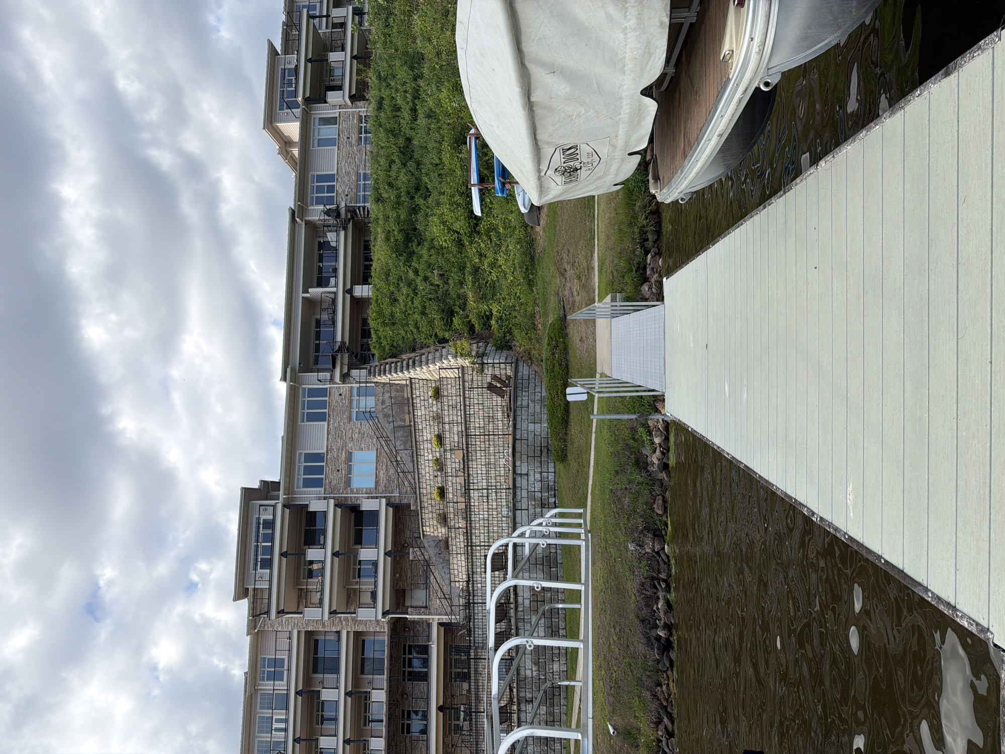 An eye-level, wide shot captures a serene lakeside scene under an overcast sky. On the left, several multi-story residential buildings, likely condominiums or apartments, rise from a sloped, grassy hillside. These buildings feature multiple windows, balconies, and a mix of stone and lighter-colored siding facades. Stone retaining walls and steps are visible leading down the incline between sections of the buildings.
In the foreground and extending from the right, a light-colored wooden or composite material dock stretches into calm, dark water, presumably a lake. A large, white covered boat, possibly a pontoon, is docked alongside the right edge of the frame. On its cover, a visible logo includes the text "SUNDOCK" and "EST. 2021" within a shield shape. Behind this boat, on the grassy bank, a rack holds at least two smaller watercraft, resembling kayaks or canoes.
The immediate shoreline consists of grass and rocks. Metal railings are present along the water's edge and near the base of the buildings, suggesting walkways or access points. The sky is filled with soft, grey clouds, indicating an overcast daytime. No people are visible, and there is no explicit activity taking place, giving the scene a peaceful and quiet ambiance. The location is consistent with a residential area on Lake Delton.