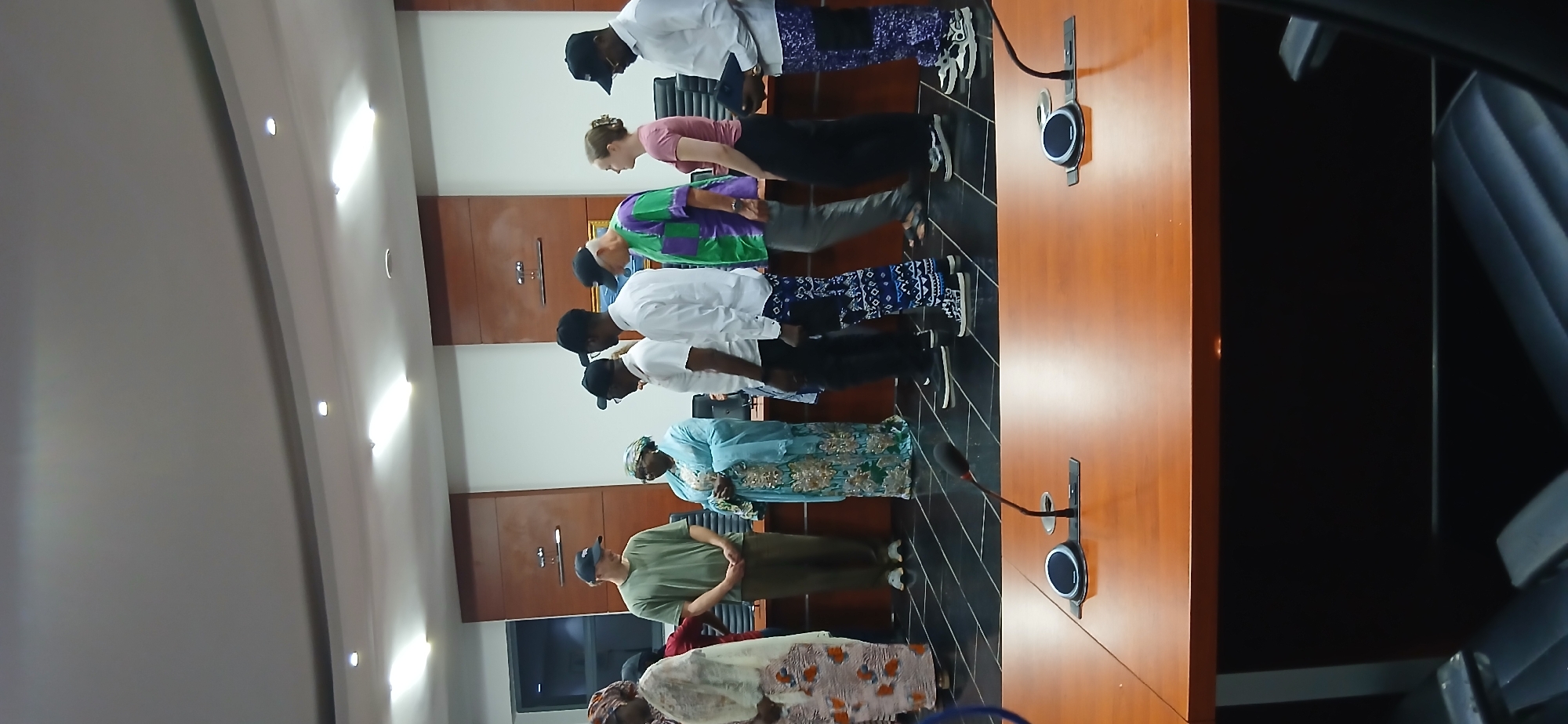 In Unguwan Sanusi, Nigeria, a group of people are standing in a line in what appears to be a conference room. The room has a long, wooden table in the foreground with two microphones on it. The walls are paneled with wood and the ceiling has recessed lighting.
The group consists of six men and two women. One woman on the far left is wearing a patterned headscarf and a long, light-colored dress with an orange and white pattern. To her right, a man in a green t-shirt and dark pants stands next to a woman in a blue and gold patterned dress and headscarf. Next to her, a man in a white shirt and dark pants with a white pattern stands with his hands behind his back. He is followed by a man in a green and purple patterned shirt and grey pants. To his right, a woman in a pink shirt and a black skirt stands next to a man in a white shirt and dark pants with a purple and white pattern. All of the men are wearing baseball caps. The people appear to be posing for a photograph or attending an event.