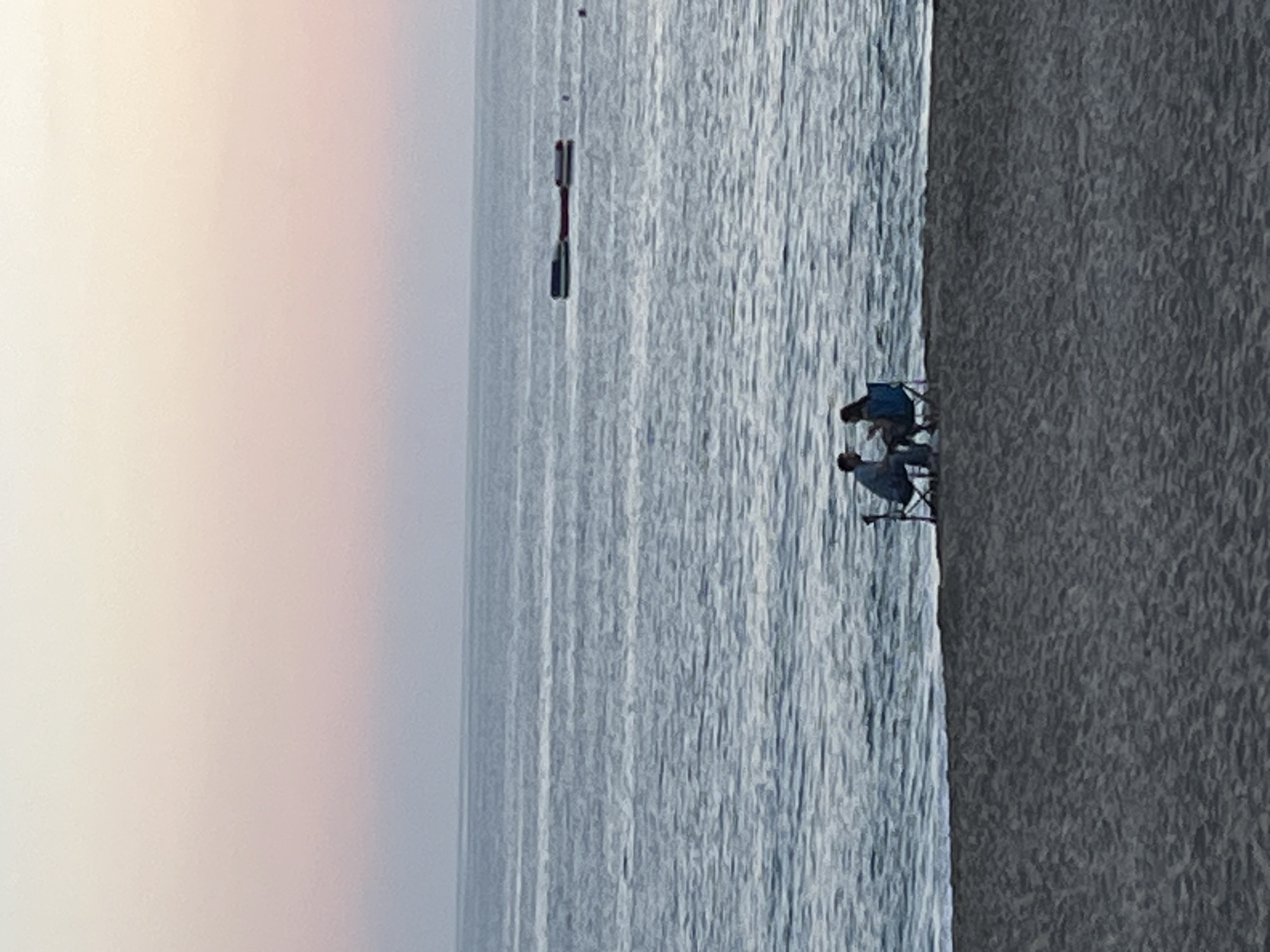 The image depicts a serene coastal scene, likely during sunrise or sunset, given the soft, diffused light and warm hues in the sky. The photograph is oriented vertically, but when rotated horizontally, it shows a wide expanse of ocean meeting a sandy beach.
The primary subjects are two individuals seated on folding chairs at the water's edge on the beach. One person is clearly visible in a blue folding chair, facing the calm sea. The second individual is partially obscured, also appearing to be seated or standing near another chair beside the first person. Their presence suggests a leisurely activity, possibly relaxing or fishing by the water.
The upper portion of the image, representing the sky, displays a gentle gradient from a pale, creamy yellow on the left, transitioning into soft pink and light blue-grey tones towards the right. This subtle coloring contributes to the tranquil atmosphere.
The majority of the image is dominated by the calm, greyish-blue water of the ocean, which shows a textured surface from gentle ripples or small waves. Further out in the water, several dark, elongated objects are visible, possibly buoys or parts of a floating structure, stretching horizontally across the middle distance.
To the right, a broad expanse of sandy beach, appearing dark grey in the current lighting, stretches along the coastline. The overall mood of the image is peaceful and contemplative, capturing a quiet moment by the sea in Alanya, Turkey. No visible text is present in the image.