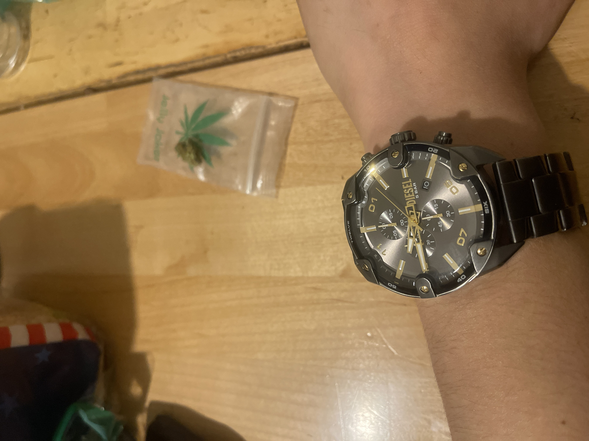 An overhead shot shows a person's wrist wearing a black Diesel watch with a gold face. The watch is on a wooden table, next to a small plastic bag containing a green bud and a marijuana leaf imprint. In the background, out of focus, is an American flag-themed object.