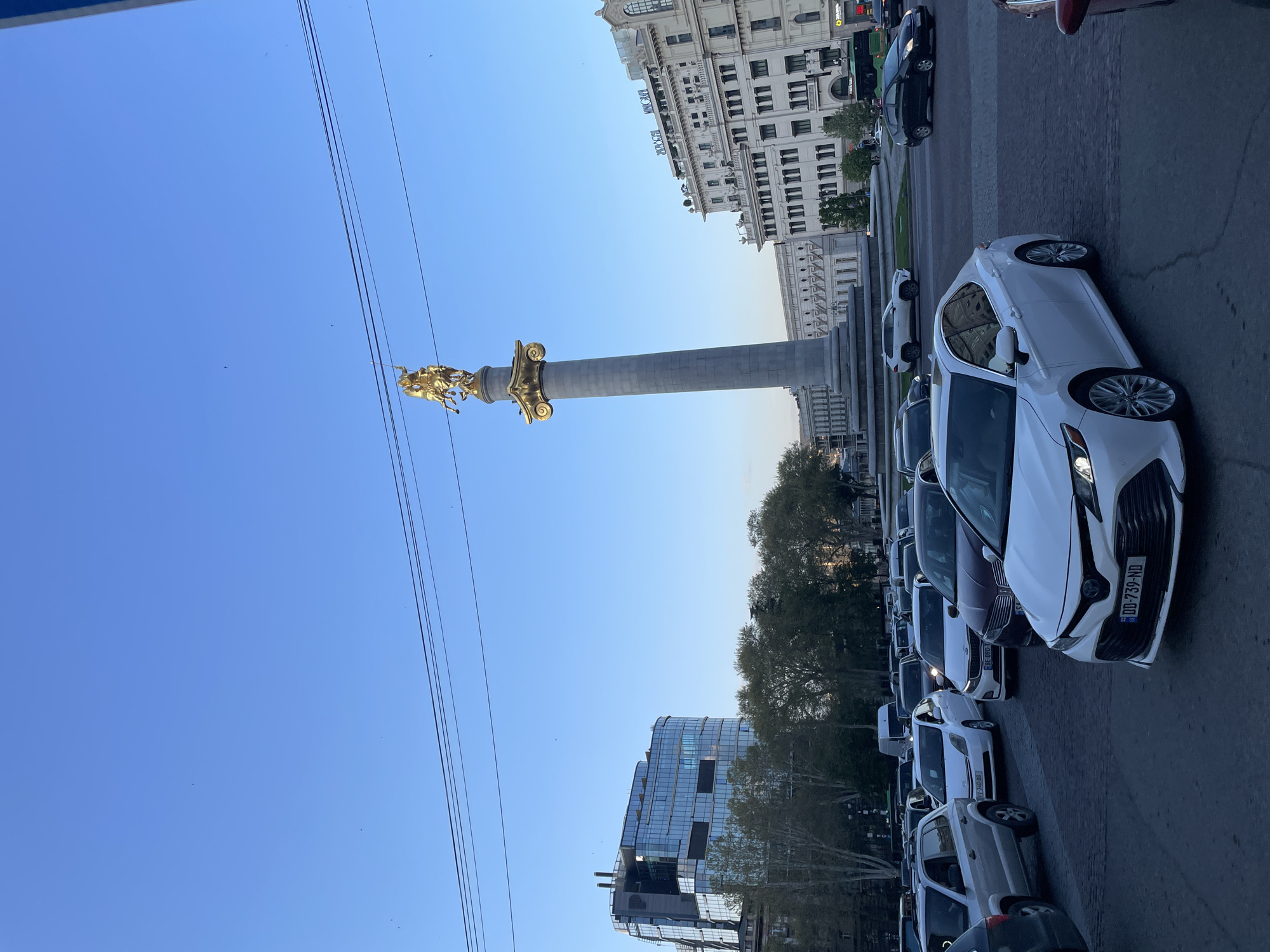 This vertical cityscape captures Liberty Square in Tbilisi, Georgia, under a clear, bright blue sky. The focal point is the towering St. George Statue, a large gilded equestrian monument depicting St. George slaying the dragon, atop a tall grey column.
The scene is set on a busy city street, with numerous cars parked in rows along the right side of the frame. A prominent white Toyota Camry sedan with the Georgian license plate "DD-739-ND" is visible in the foreground. Other white and dark colored sedans, some with visible Georgian plates like "BE-109-TB" and "MV-540-HB", extend into the background, suggesting a bustling urban area.
To the left of the monument, a large, ornate multi-story building with classical architectural elements stands, bearing the visible text "RUSTAVELI" and "TBC BANK" on different sections. Further in the background, a modern glass-clad building is partially obscured by trees. Power lines crisscross the upper left portion of the sky.
The lighting suggests either late afternoon or early morning, with bright sunlight illuminating the gilded statue and parts of the buildings, casting soft shadows. The weather is clear and sunny, indicating fair conditions. The absence of people in the immediate foreground shifts the focus to the urban environment and its architectural and monumental landmarks.
