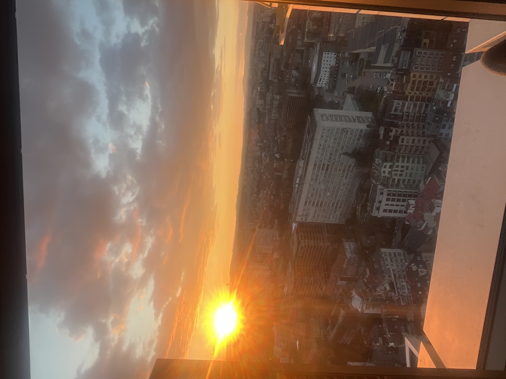 The image captures a sunset over Sofia, Bulgaria, from a high vantage point, likely within a building. The sky is dominated by orange and yellow hues, with clouds illuminated by the setting sun. Below, the city unfolds with its mix of buildings, ranging from low-rise structures to high-rise apartment complexes. Major structures include a large, white building with numerous windows, suggesting a commercial or residential complex. Street grids are visible, revealing traffic patterns and urban planning. The view is partially framed by the building's window frame.