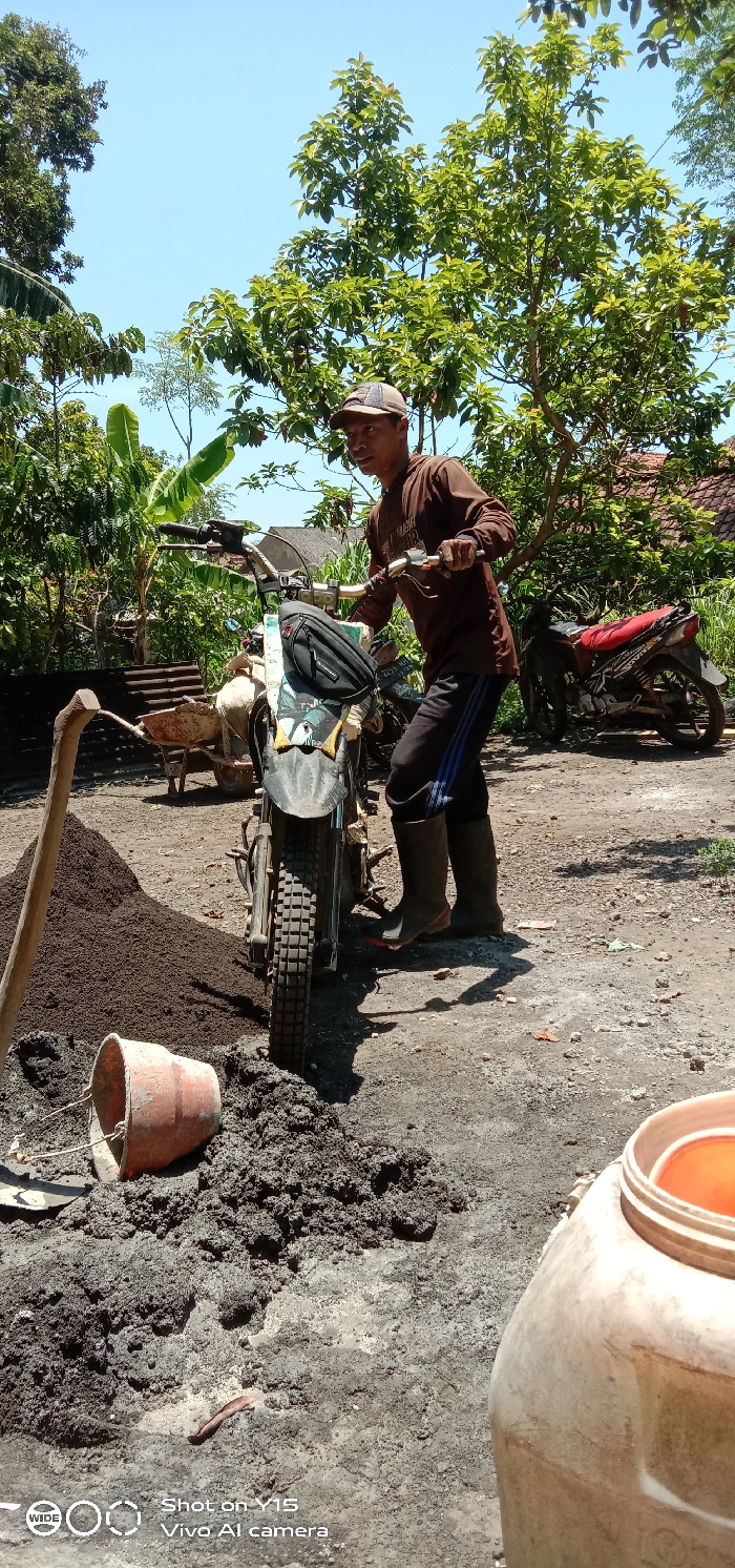 This image captures an outdoor construction or gardening scene in Ranugedang, Indonesia, under bright daylight. In the foreground, a male individual, appearing to be an adult, is standing to the left of a dirt bike-style motorcycle, holding its handlebars. He wears a brown long-sleeved shirt, dark blue track pants with light blue stripes, a light-colored cap, and dark boots. His gaze is directed slightly to his right, with a subtle smile or grimace on his face.
The motorcycle is positioned centrally, featuring knobby tires and a black bag strapped to its handlebars. To the left of the motorcycle, a significant pile of dark brown, granular material (likely soil or sand) is visible, with a wooden-handled hoe leaning into it. Immediately in front of the motorcycle and to the right of the granular pile, a substantial heap of wet, dark grey, mixed material (resembling cement or mortar) lies on the ground. An orange plastic bucket is on its side next to this mixture. In the bottom right corner, a white plastic jerrycan with an orange lid is partially visible.
In the mid-ground, behind the granular pile, a red wheelbarrow with wooden handles is present. Further back on the right, another motorcycle with a red seat and dark body is parked. The background is dominated by lush green foliage, including tall trees and banana plants, under a clear blue sky. The ground throughout the scene is uneven, unpaved dirt with scattered debris, indicative of a working area. Shadows are cast by the man and objects, suggesting strong overhead sunlight.