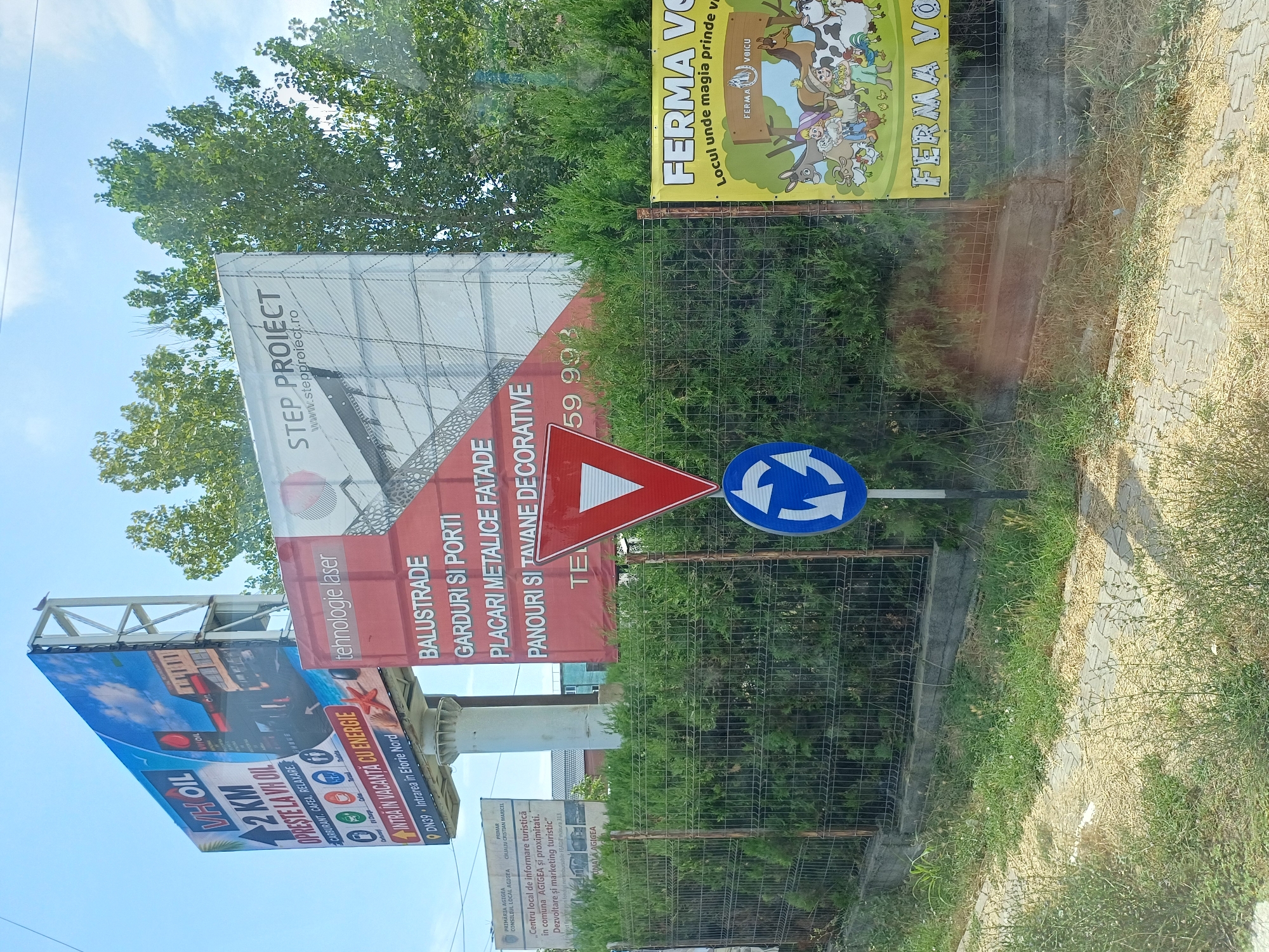 The image captures a scene in Agigea, Romania, featuring several billboards and traffic signs along a roadside. The primary focus is on large advertisements, including one for "VH OIL" indicating a fuel station with amenities, another for "STEP PROJECT" advertising laser technology, and a red billboard listing various construction materials. A yellow banner on the right side advertises "FERMA VOICU", a farm where magic comes alive, depicting cartoon farm animals and people. Traffic signs, a yield sign (inverted red triangle), and a roundabout sign (blue circle with arrows) are also prominently displayed. The environment is characterized by lush green bushes and trees, a wire fence, and a paved roadside with patchy grass. The sky is bright and appears to be partly cloudy. There are no people clearly visible in the scene.