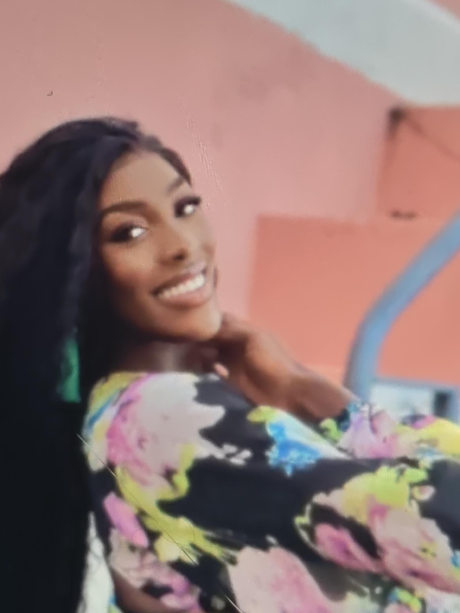 A medium shot captures a young woman with dark skin and dark hair, smiling with her teeth showing. She is wearing a dark, long-sleeved shirt with a floral pattern featuring pink, yellow, and blue flowers. Her head is tilted to the side, and her right hand is resting on her neck. The background is a blurry pink wall with a light-colored architectural element on the right. The image appears to be taken indoors.