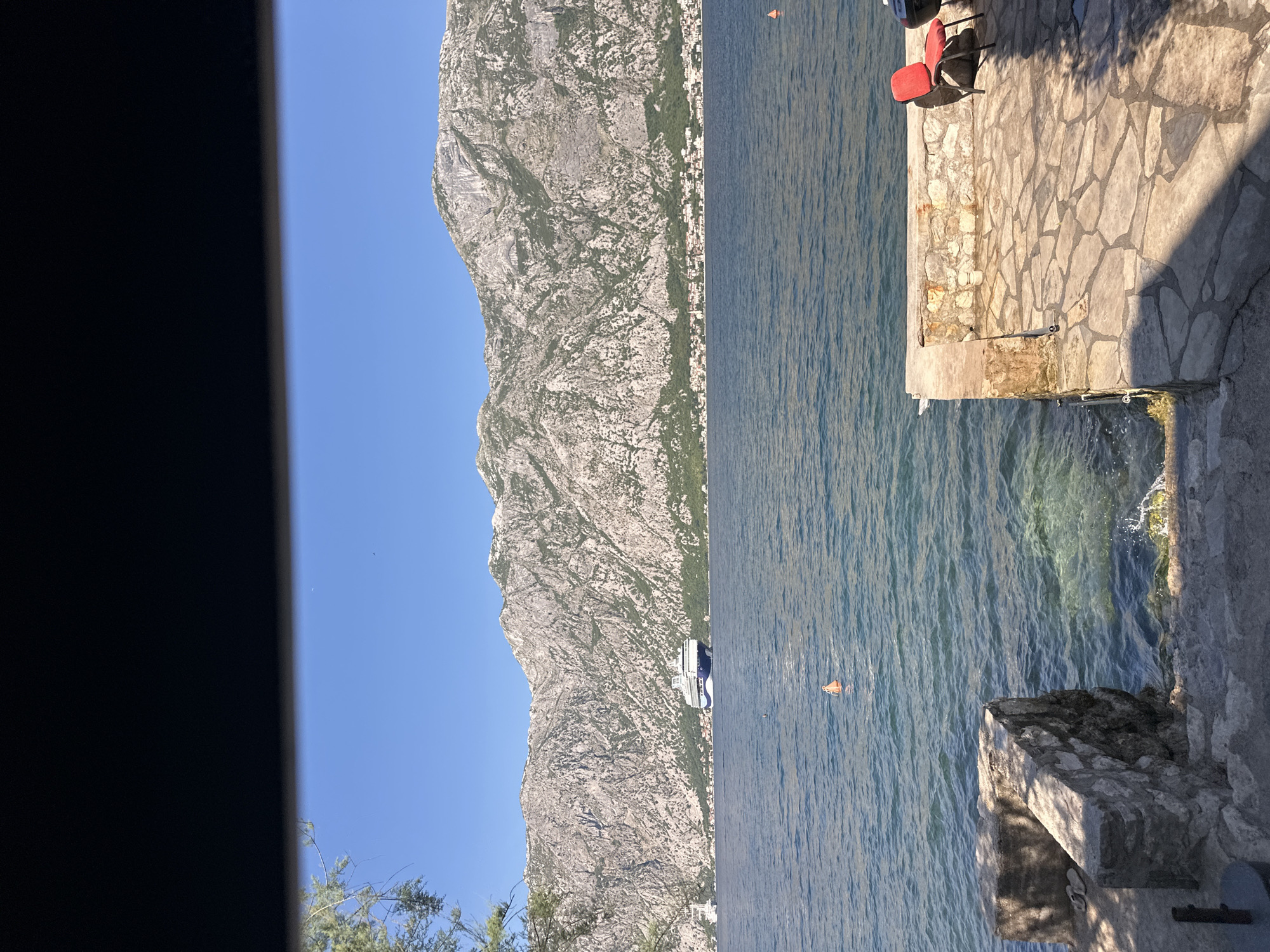 The image depicts a scenic view of the Bay of Kotor, near the town of Prčanj, Montenegro. In the foreground, a stone-paved patio area features a red chair, casting a shadow on the stones. To the left, stone steps lead down towards the clear blue water, with a wall constructed of large stones. The water itself is calm, with a few buoys visible. In the middle ground, a large white and blue cruise ship is anchored in the bay. The background is dominated by the imposing, rocky mountains that frame the bay, with a cluster of buildings visible along the shoreline at the base of the mountains. The sky is a clear, bright blue, indicating a sunny day. A dark, horizontal element is visible at the very top of the image, suggesting it was taken from under an overhang or shelter.