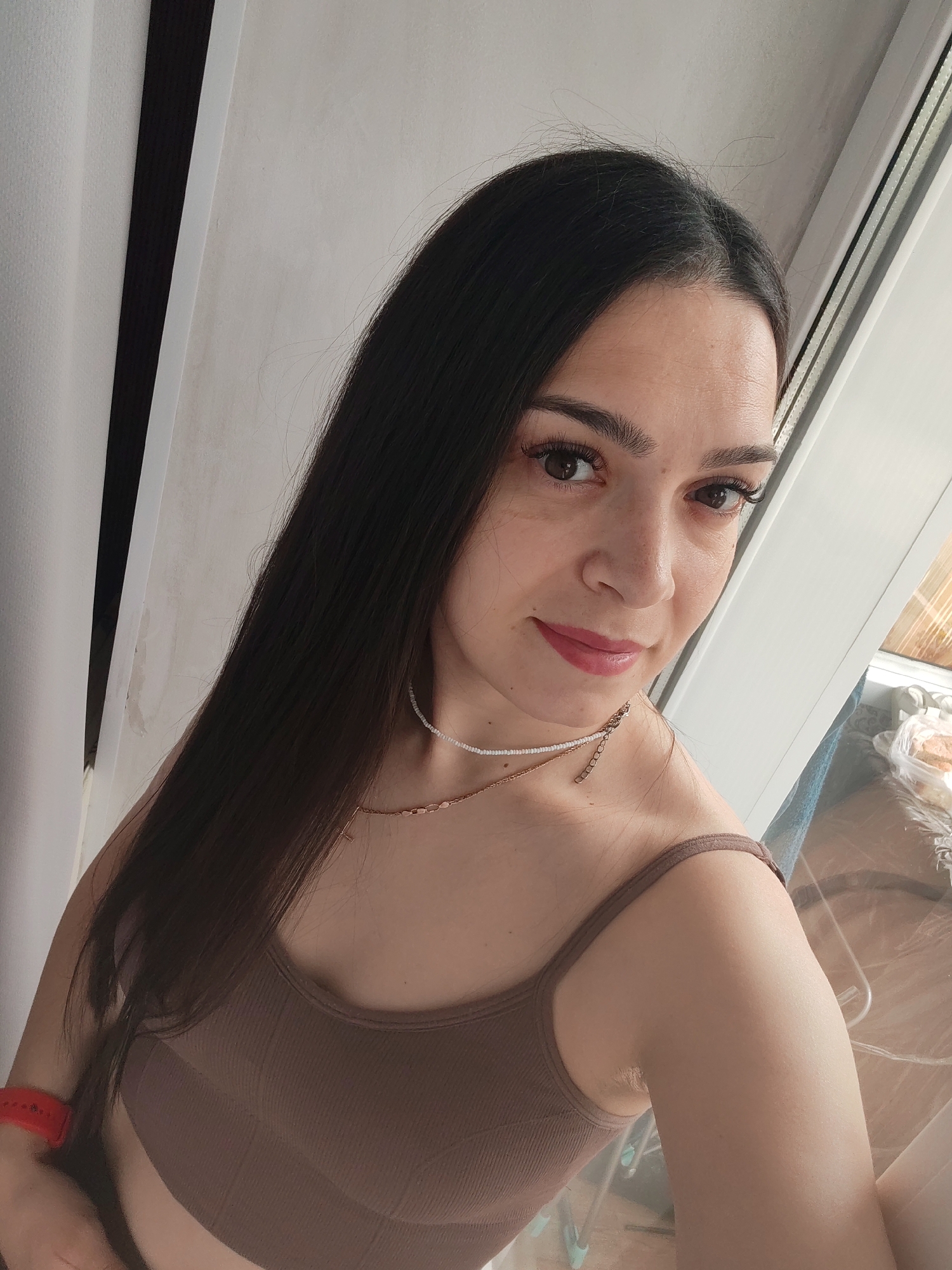 A young woman with long, dark hair is taking a selfie indoors. She is wearing a brown tank top and two necklaces. Her right arm is bent, and she is wearing a red fitness tracker. The background is a plain wall with vertical blinds. A window is visible to the right, showing some objects in a plastic bag. The lighting suggests it is daytime. There are no indications of the location being Belogorsk, Russia.