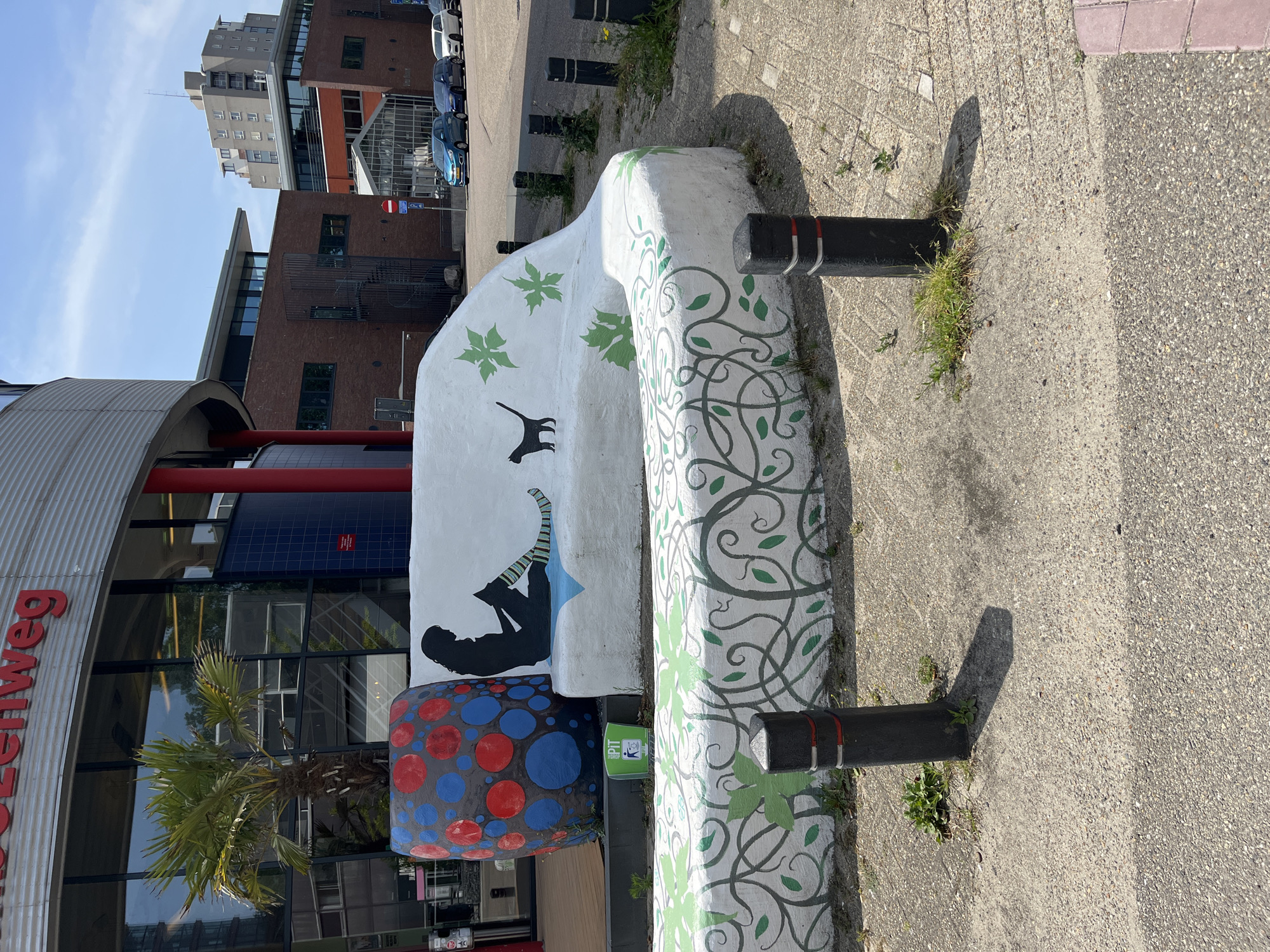 An outdoor medium shot captures a curved white bench adorned with green ivy-like artwork and a silhouette of a person reading. To the left of the bench, a large, colorful planter with red and blue dots holds a small palm tree. In the background, a modern brick building with a glass-enclosed section and a multi-story building are visible, along with several parked cars on a paved street. The bench is situated on a patch of sand and gravel, bordered by a paved walkway. The sky is clear and blue.
