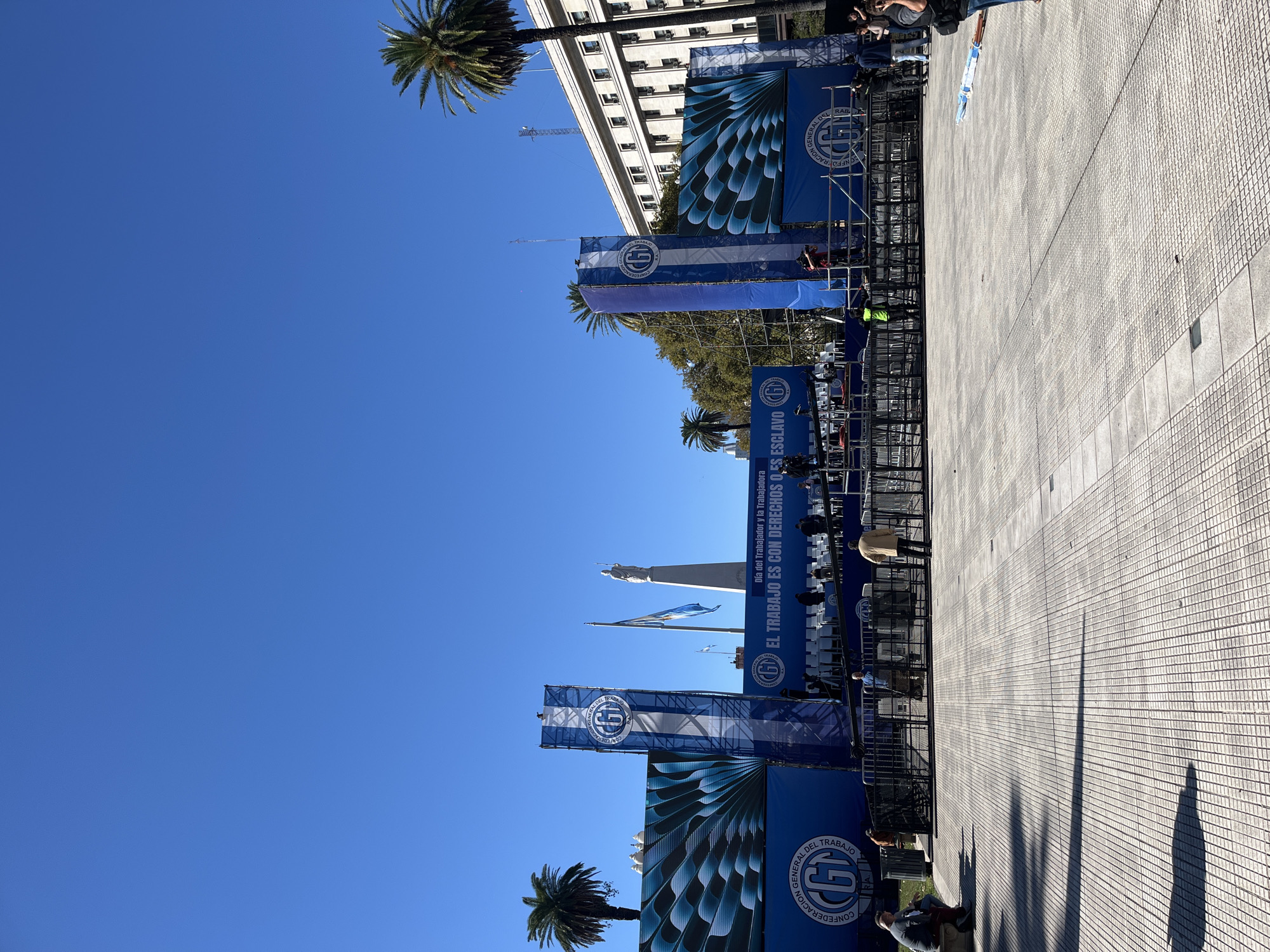 This media file captures an outdoor public square in Buenos Aires, Argentina, on a bright, sunny day with a clear blue sky. The scene is set up for a public event, likely a rally or celebration centered around labor rights.
Numerous large blue banners, supported by metal scaffolding, dominate the foreground and midground. These banners prominently display the logo and full name of the "CONFEDERACIÓN GENERAL DEL TRABAJO" (General Confederation of Labor). One notable banner carries the slogan "EL TRABAJO ES CON DERECHOS O ES ESCLAVO" (Work is with rights or it is slavery), while another mentions "Día del Trabajador y la Trabajadora" (Worker's Day). The logo features a white stylized 'G' within a circle, set against a blue and white striped background on some banners.
Several people are visible throughout the square; some are standing near the banners and structures, while others are seated on white chairs arranged in rows, suggesting an audience or participants for the event. Palm trees are scattered around, adding to the outdoor setting. In the background, a large, ornate white building with many windows and balconies stands, possibly a government edifice. A tall, white monument topped with a statue can also be seen further in the distance.
The ground is a vast, paved area with a grid pattern, typical of urban plazas. The strong, defined shadows cast by the structures and individuals indicate that it is midday or early afternoon. The overall atmosphere suggests an organized public gathering with a clear message related to workers' rights.