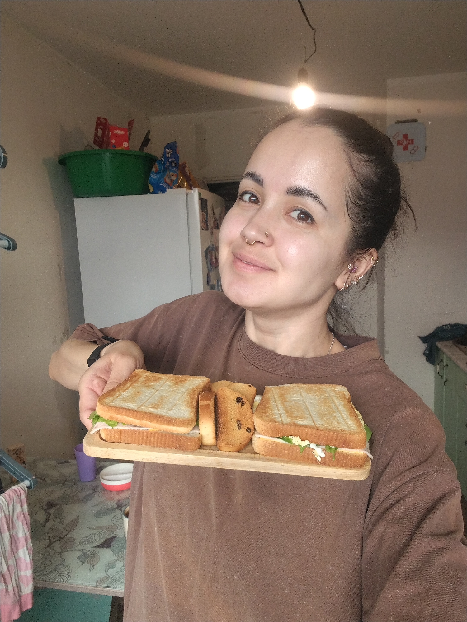A person is holding a wooden tray with two sandwiches and a piece of raisin toast. The sandwiches appear to be made with toasted bread, lettuce, and a filling, possibly egg salad. The person is wearing a brown long-sleeved shirt and has their hair pulled back. They have multiple ear piercings and a nose ring. The background shows a kitchen with a refrigerator, various containers on top of it, and a first-aid kit on the wall. A bare light bulb hangs from the ceiling. It is not possible to verify the location in Noviy, Russia based on the visual information provided.