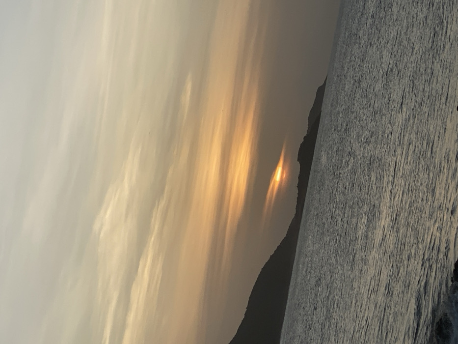 A hazy sunset over the ocean. Light glares through the atmosphere, creating streaks of bright orange against a light, cloudy sky. The sun, partially obscured by a dark, silhouetted coastline, reflects on the water. The ocean appears rippled and textured, transitioning from greyish-blue to darker shades towards the bottom of the image.