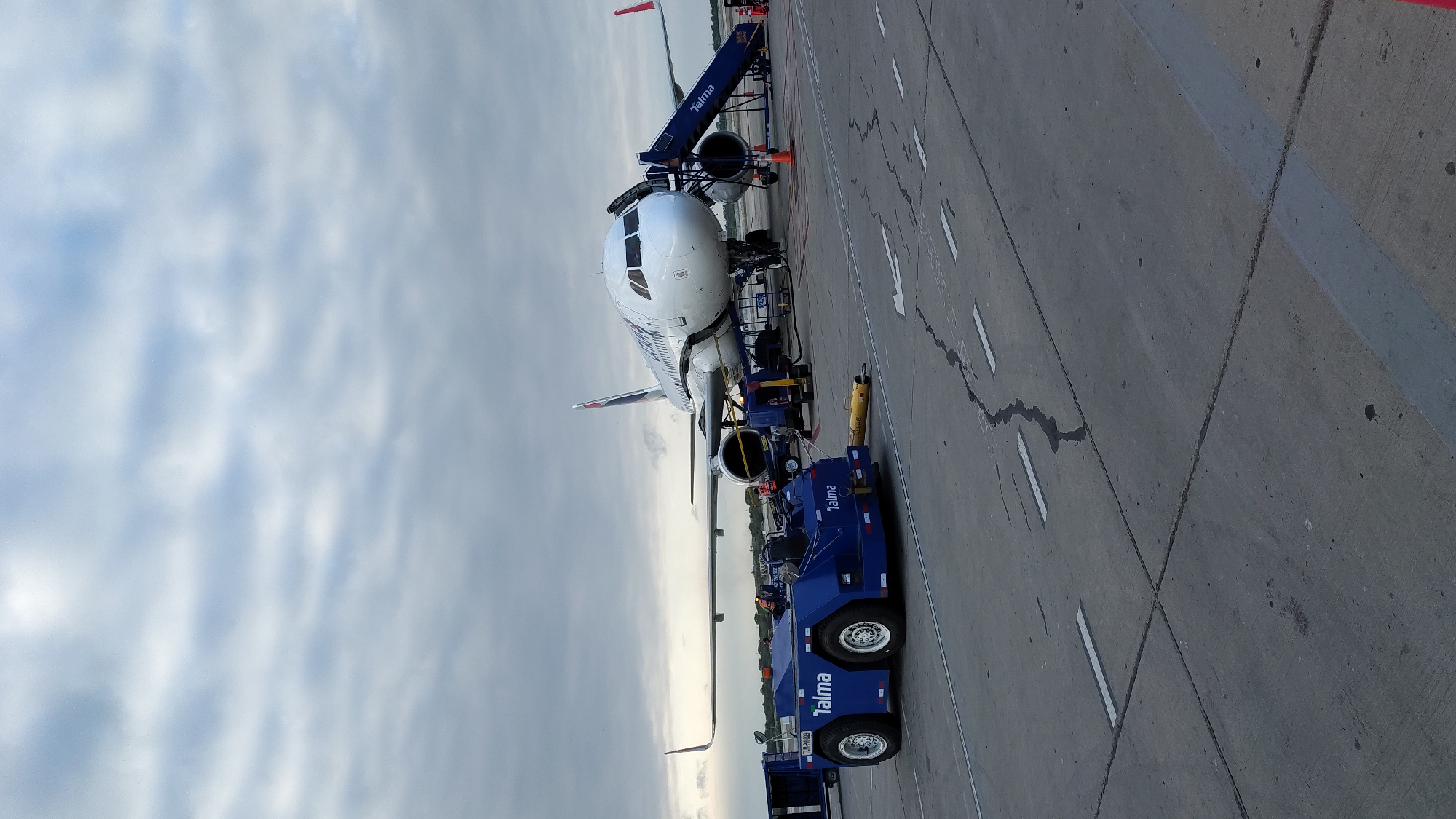 The image depicts an airplane parked on a tarmac. A blue "Talma" pushback truck is attached to the aircraft's front. A boarding stair is connected to the front of the airplane. The sky is visible in the background. The location cannot be determined.