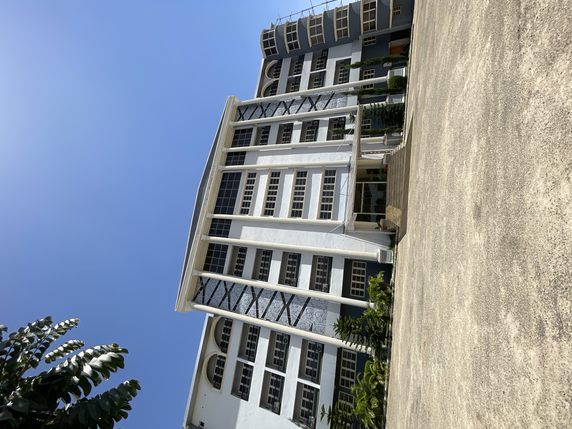 The image shows a tall, multi-story building in Jos, Nigeria, under a clear blue sky. The building has a modern architectural design, with multiple rows of windows and decorative columns. There's a textured concrete surface in the foreground, likely a walkway or parking area. Green foliage is visible near the base of the building and in the lower left corner of the image.