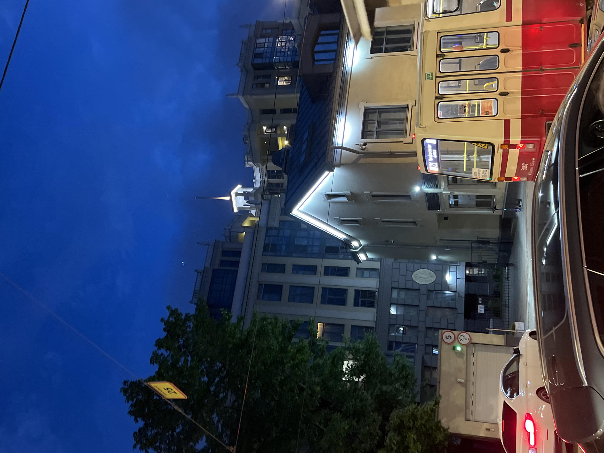 The image depicts a twilight scene in Saint Petersburg, Russia, likely from a street-level perspective. Dominating the foreground is the side of a vintage tram, painted in cream and red, with its doors visible and illuminated interior. The tram is numbered 5087 and displays a route number 21 and destination sign in Cyrillic. To the left of the tram, a white car with its taillights illuminated is parked or stopped. Behind the tram and car, a series of multi-story buildings are visible. A lower, cream-colored building with illuminated windows and roofline lights is directly behind the tram. Further back, a taller, modern apartment building with a lighter facade and numerous windows is prominent. The sky is a deep blue, indicating the late hour. A large green tree with dark foliage is situated to the left of the frame, partially obscuring the buildings. A yellow street sign with the number '25' is suspended from a wire above the tree. Additional street signs, including a speed limit indicator and a height restriction sign, are visible on a pole near the white car. The overall environment suggests an urban setting with street infrastructure and residential or commercial buildings.