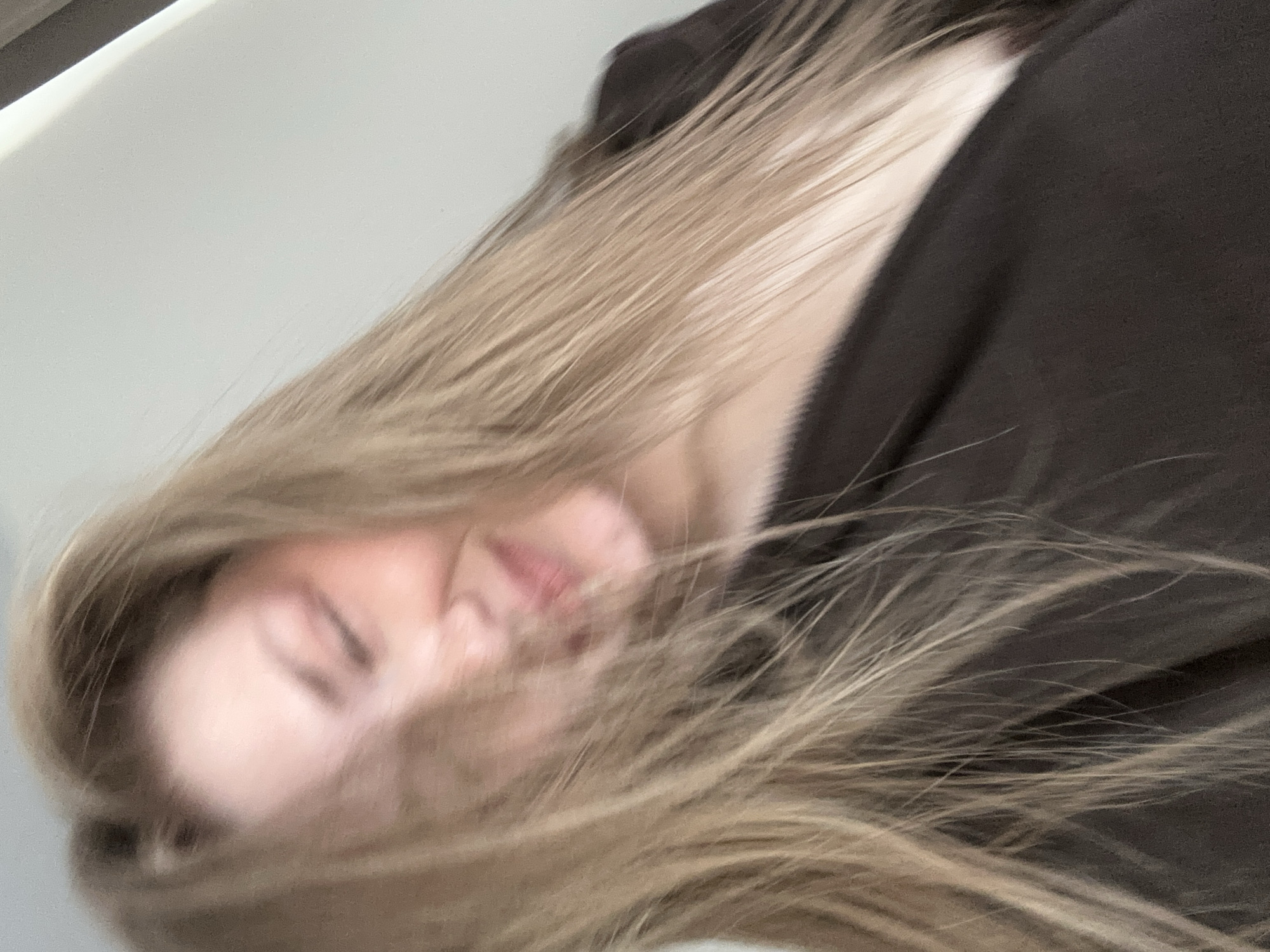 The image displays a close-up, angled portrait of a single individual, likely female, situated in Kazan, Russia. The subject's face is partially obscured by long, light brown hair, which appears to be in motion. Observable facial features include fair skin with a slight blush, eyes that are closed or squinting, and a discernible smile on the blurred lips. The individual is wearing a dark brown garment with an open neckline, revealing a portion of the chest. The background is uniformly light-colored and indistinct, suggesting an indoor environment such as a wall or ceiling.