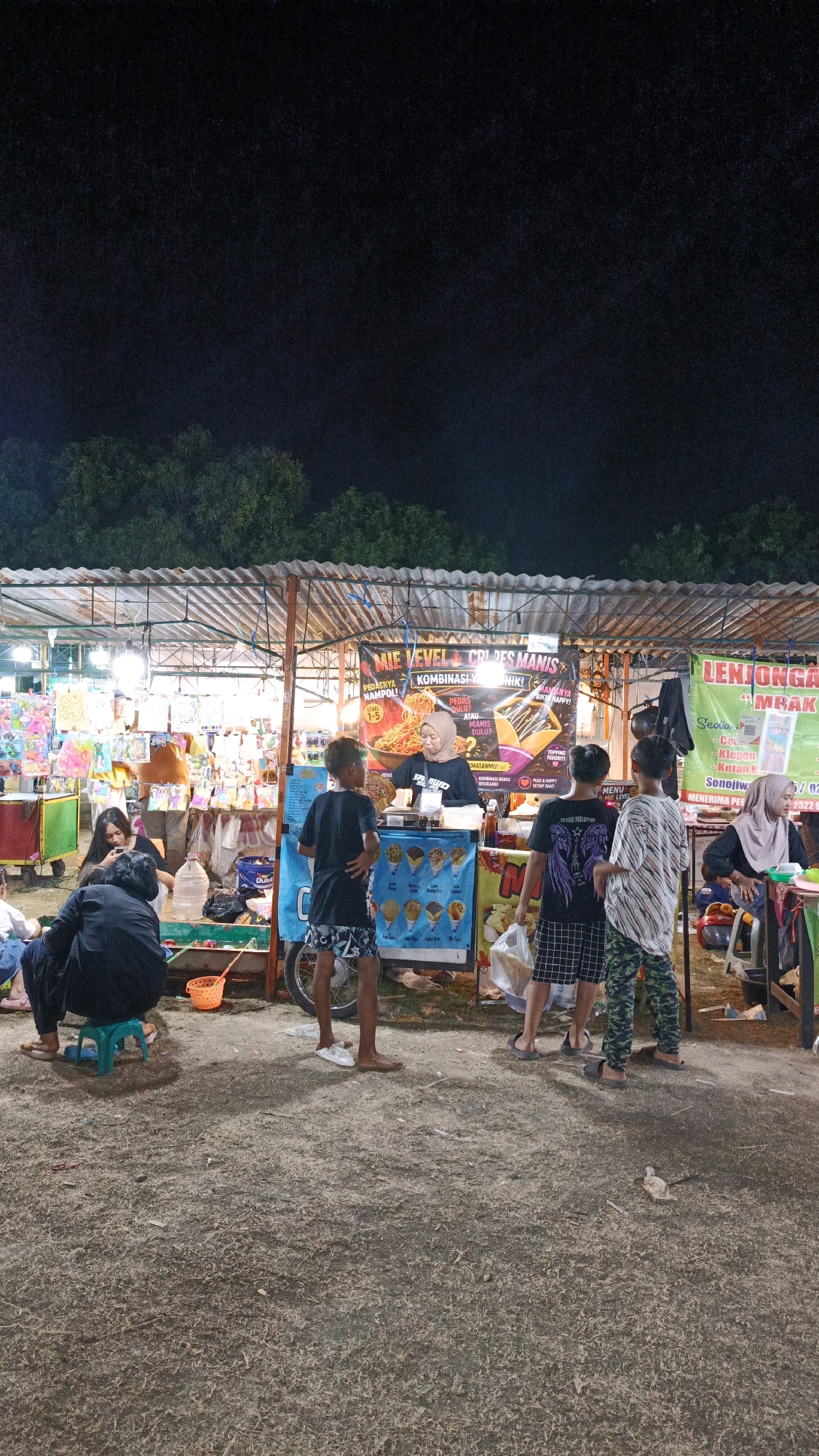This image captures a nighttime scene at a street food stall in Surakarta, Indonesia. The stall appears to be part of a market or festival, with several people gathered around.
In the foreground, a group of children and young adults are standing and sitting. Three young boys are prominently positioned in front of the stall, looking towards the vendor. One boy on the left wears a black t-shirt and patterned shorts, another in the center has a black t-shirt with a graphic design and plaid shorts, and the boy on the right is in a striped t-shirt and camouflage-patterned pants. To the left, a young person in a black outfit sits on a small green stool, while another person in dark clothing sits beside them.
The stall itself is covered by a corrugated metal roof. A banner above the stall advertises "MIE VEVEL & CREPES MANIS" (Noodle Vevel & Sweet Crepes) and "LENJONGAK MBAK" (A type of snack). A female vendor, wearing a hijab and a dark t-shirt, stands behind the counter, preparing food. In front of the stall is a display of ice cream cones. Various food items, drinks, and products are visible on shelves and displays within the stall. A large blue water jug is placed near the seating area on the left.
The ground is a mix of dirt and dry grass, with some litter scattered around. Trees are visible in the background, illuminated by the artificial lights of the market. The overall atmosphere suggests a lively, casual evening event.