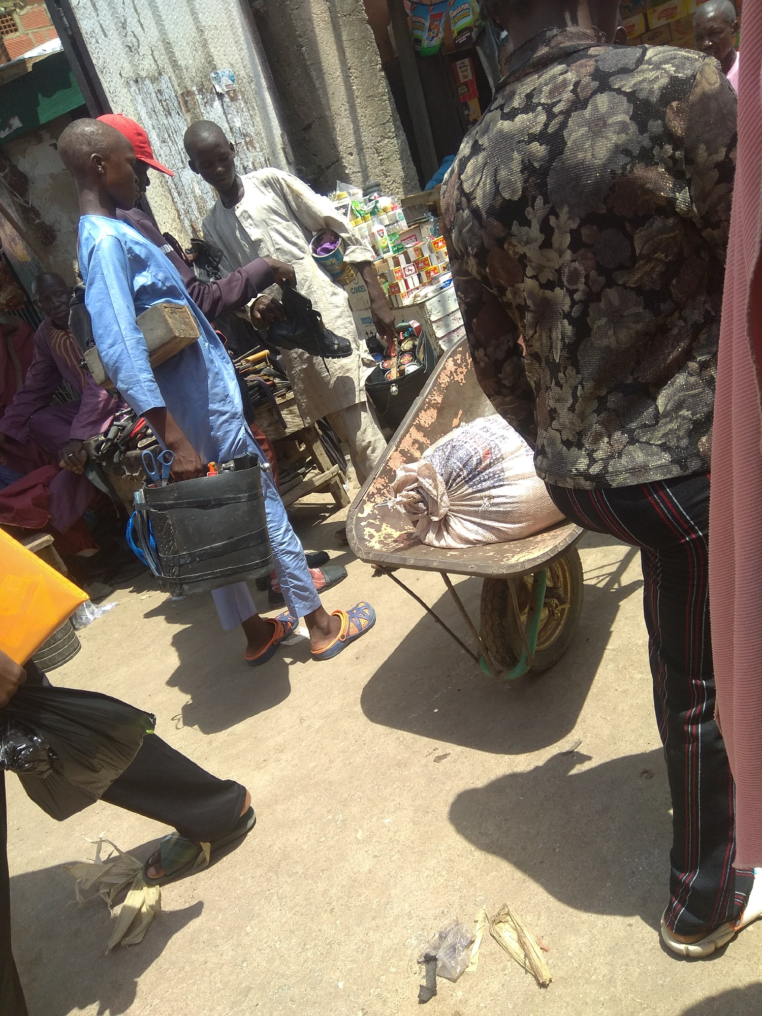 The image depicts a street scene in Jalingo, Nigeria, likely during the daytime given the strong shadows cast. The setting appears to be an outdoor market or a busy street with various people and objects.
The central focus is on a group of people interacting. One individual, wearing a blue garment and a red cap, is holding a black container with scissors protruding from it, and a wooden block is placed on their shoulder. Another person in a light-colored robe is reaching towards them, possibly showing or handing something. To the right, the back of a person wearing a dark, floral-patterned shirt and striped pants is visible, standing near a wheelbarrow. The wheelbarrow contains a large, tied sack of what appears to be grain or produce. Further in the background, other individuals and stalls can be glimpsed, suggesting a bustling environment.
The ground is a dusty, sandy surface typical of outdoor African markets. The presence of goods on display and people engaged in commerce implies a marketplace atmosphere. The overall mood seems to be one of daily activity and interaction. There is no discernible text in the image.
