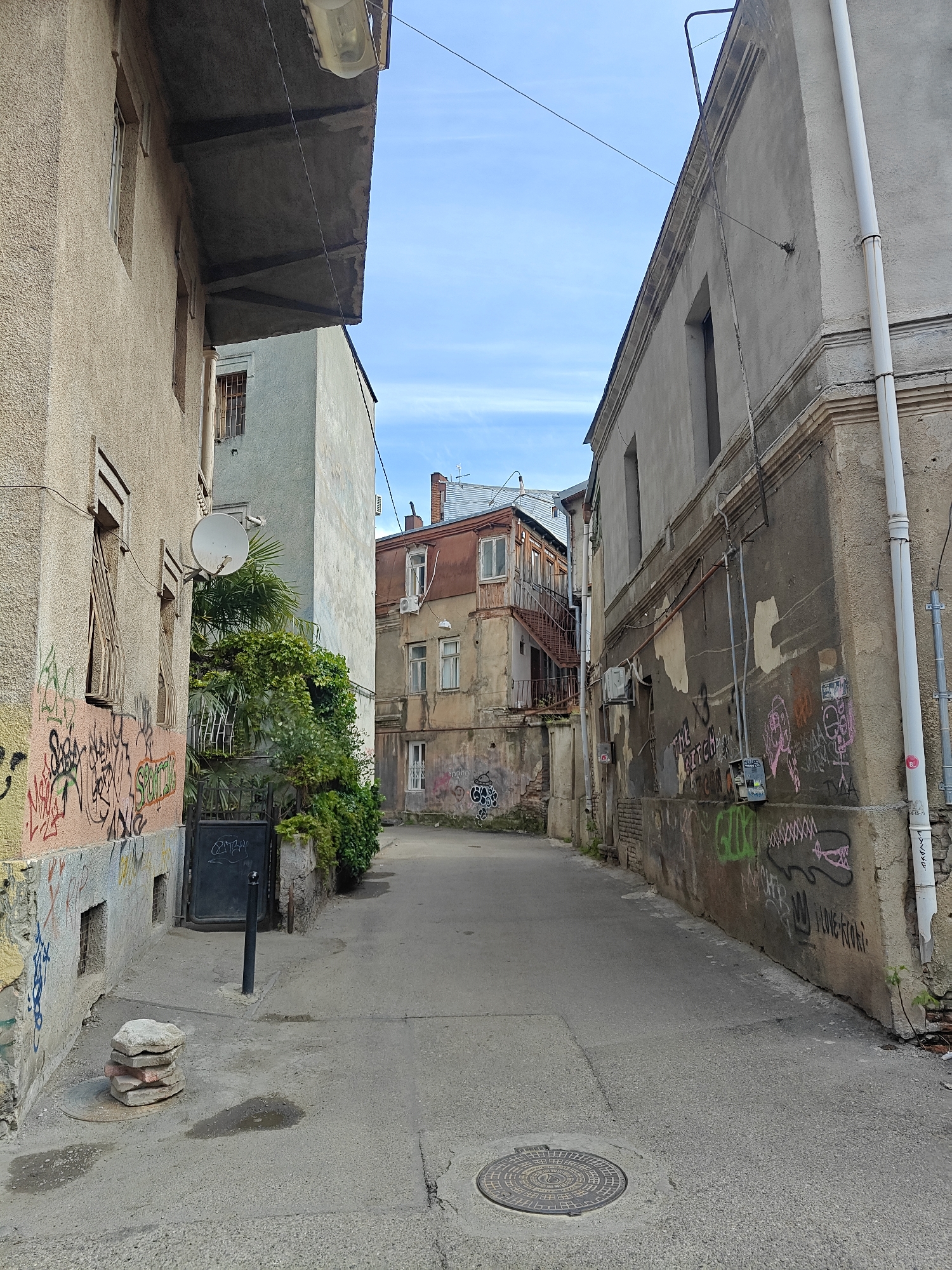 The image depicts a narrow, weathered urban alleyway in Tbilisi, Georgia, under a clear blue sky during bright daylight. The street is empty of people or vehicles.
Buildings flank both sides of the asphalt-paved alley. On the left, a light tan, textured stucco building with several windows, some barred, is heavily covered in colorful graffiti including "Saba" and "SPOW." A white satellite dish is mounted on its wall, partially obscured by green foliage, including a palm tree, which grows between the buildings. An overhanging eave with a fluorescent light fixture extends from the top of this building.
On the right, a darker gray-brown building with peeling paint and exposed concrete is also extensively covered in graffiti. Visible tags include "THE BITCH" (partially obscured), "GLLO," and "LOVE KOKI," along with a cartoonish pink and black alligator-like drawing. A white drainpipe runs vertically along its right edge, and electrical conduits and meters are attached to the wall.
Further down the alley, a light-colored, seemingly older building with a red-tiled roof, an external wooden balcony, and an external metal staircase is visible. Lush green ivy and other plants grow along the walls and behind a dark metal gate in the middle ground.
On the asphalt ground, a circular metal manhole cover is present in the foreground on the right. On the left, near the curb, a stack of flat, weathered stones or bricks sits beside a black metal bollard. Two dark, damp patches are visible on the asphalt surface. The overall impression is one of an old, lived-in urban space with a rich patina of age and human activity, notably street art.